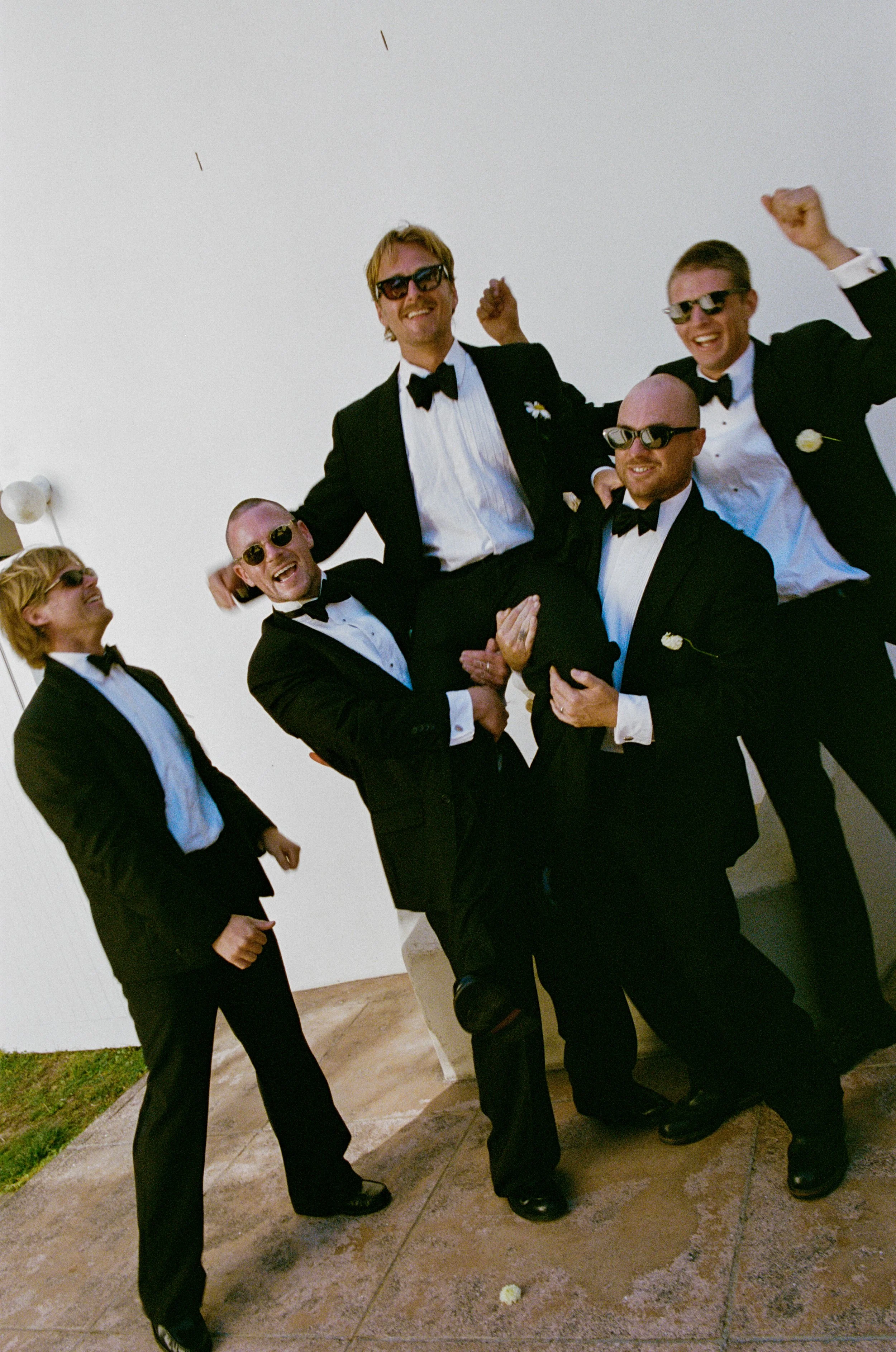 Group of six men in tuxedos and sunglasses celebrating, with one man being lifted by others, against a plain white wall.
