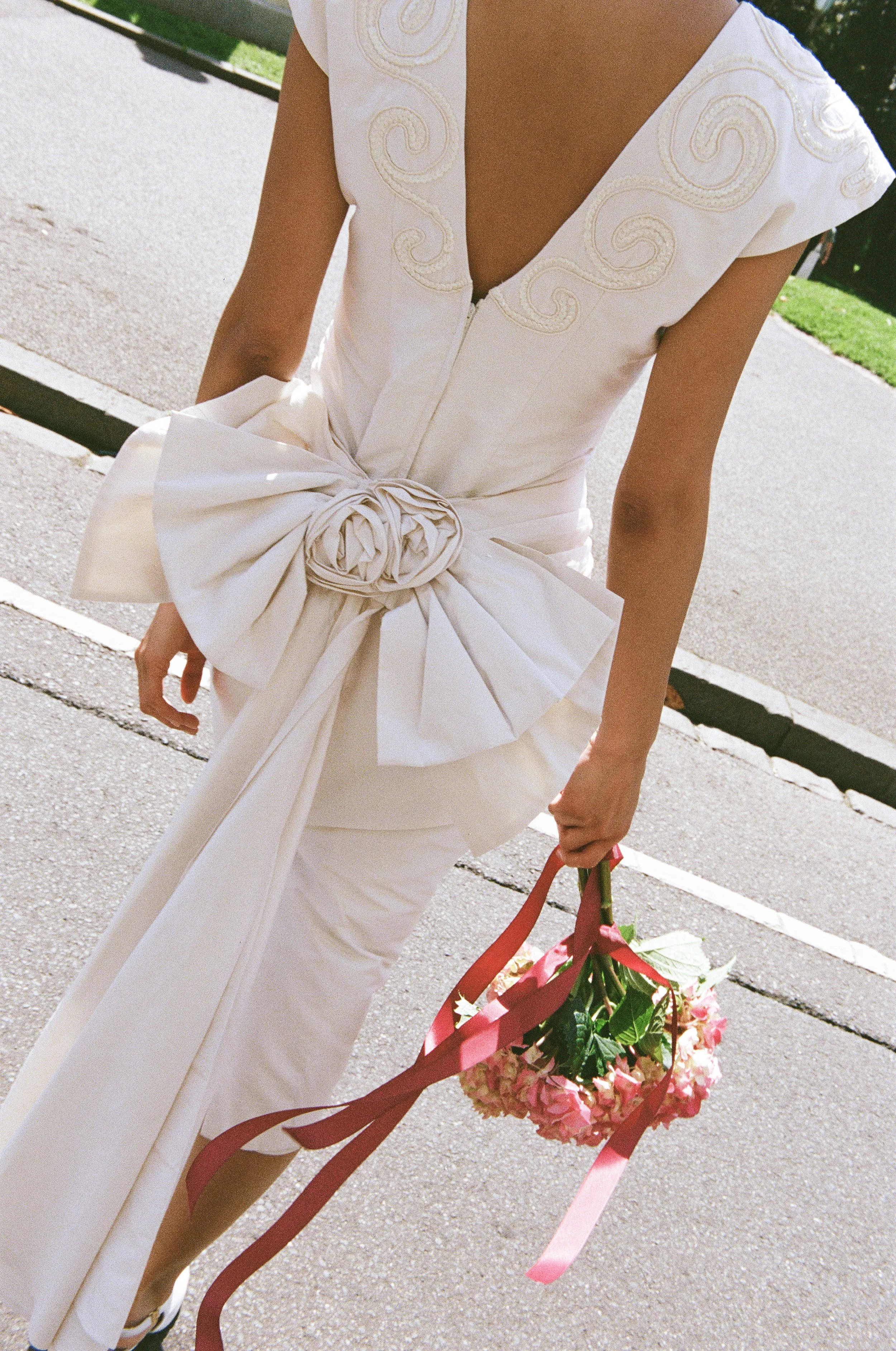 A woman in a white dress with intricate embroidery and a large bow at the waist, holding a small bouquet of pink and white flowers with pink ribbons, walking on a sidewalk.