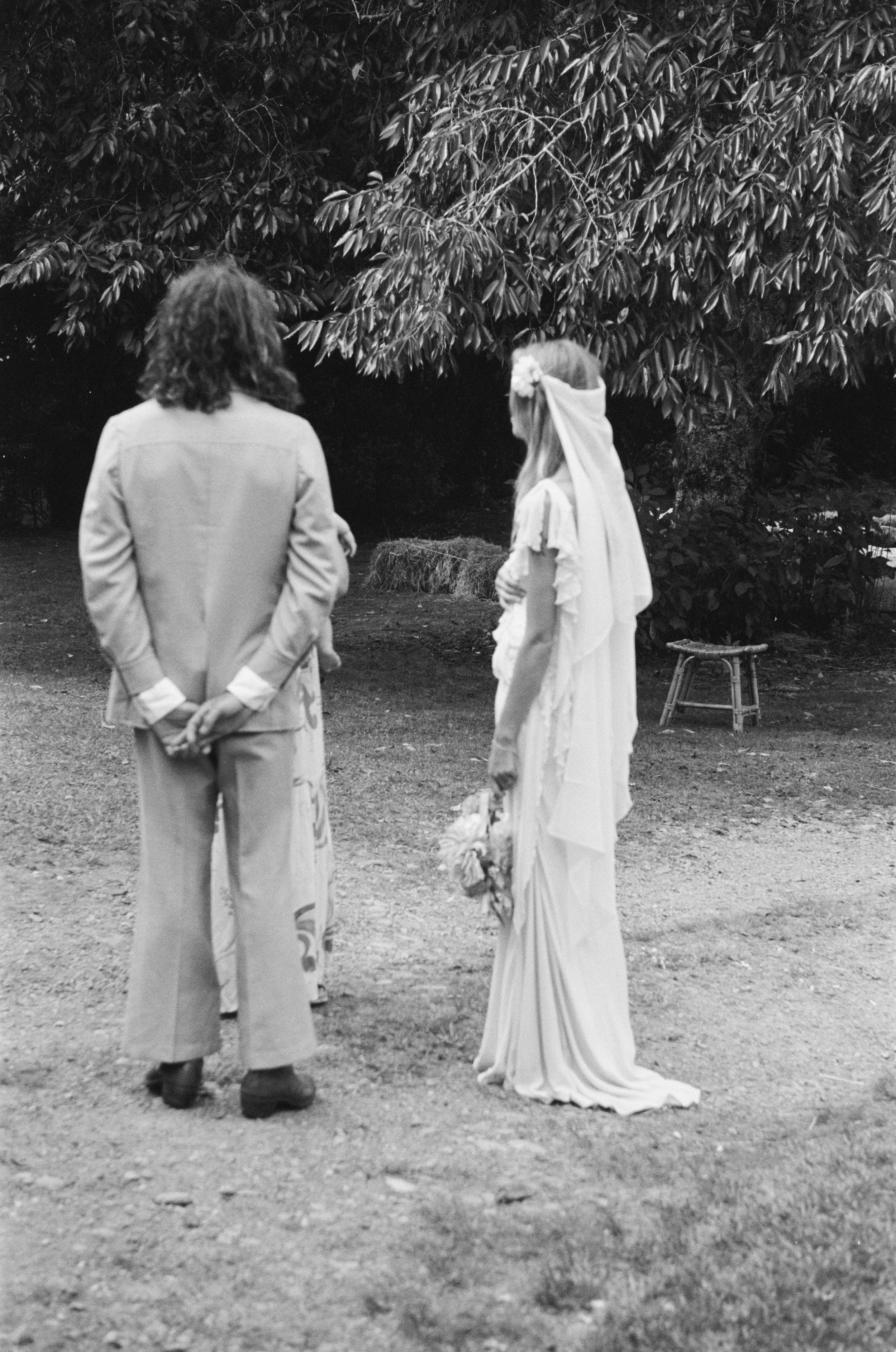 A black-and-white photo of a man and a woman standing outdoors, facing each other, with large trees in the background. The woman is dressed in a long, flowing gown with a flower in her hair and a veil. The man is dressed in casual attire with a jacket and pants, standing with his hands behind his back.