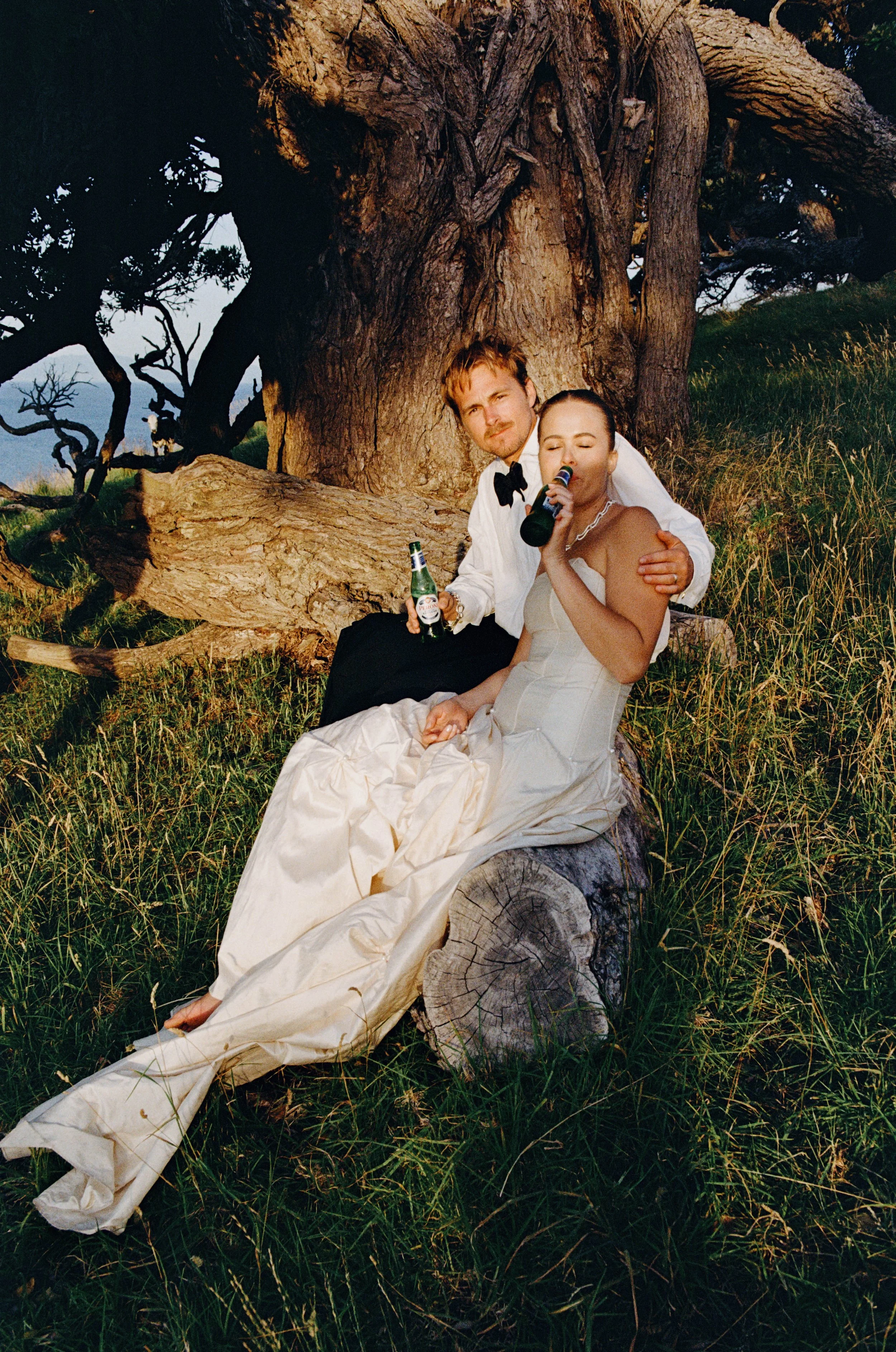 A bride and groom sitting on a log in a grassy field by a large tree, holding beers and taking a photo together.