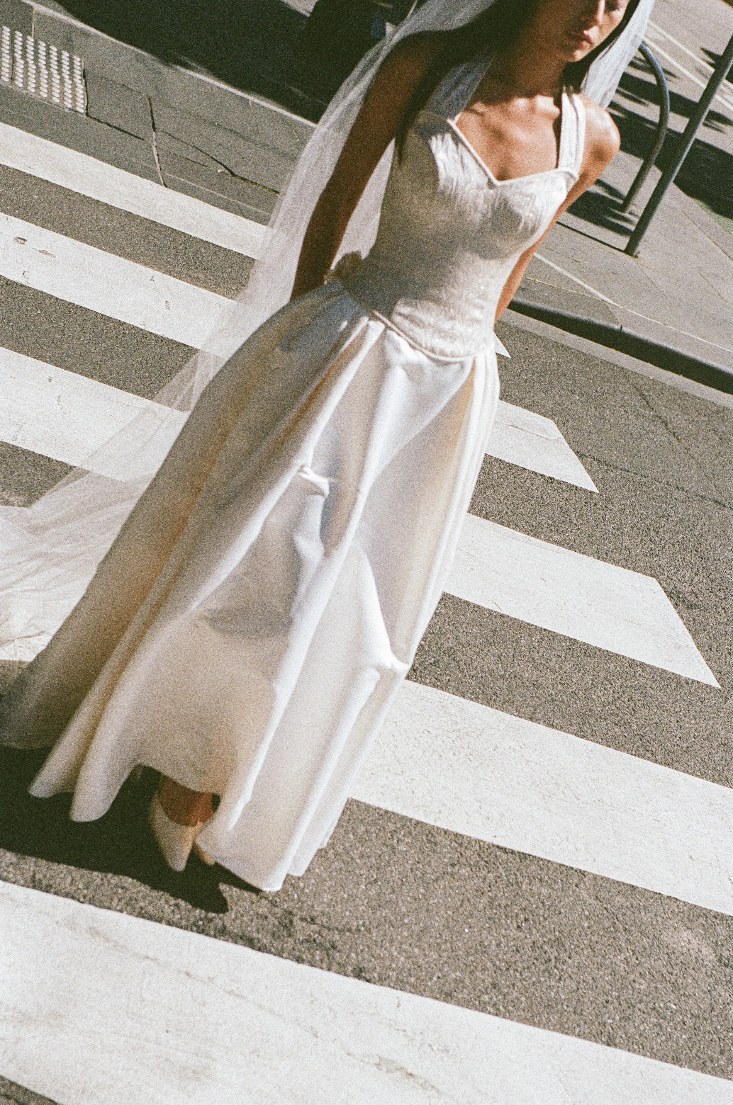 A woman dressed in a white wedding gown standing on a crosswalk on a sunny day.