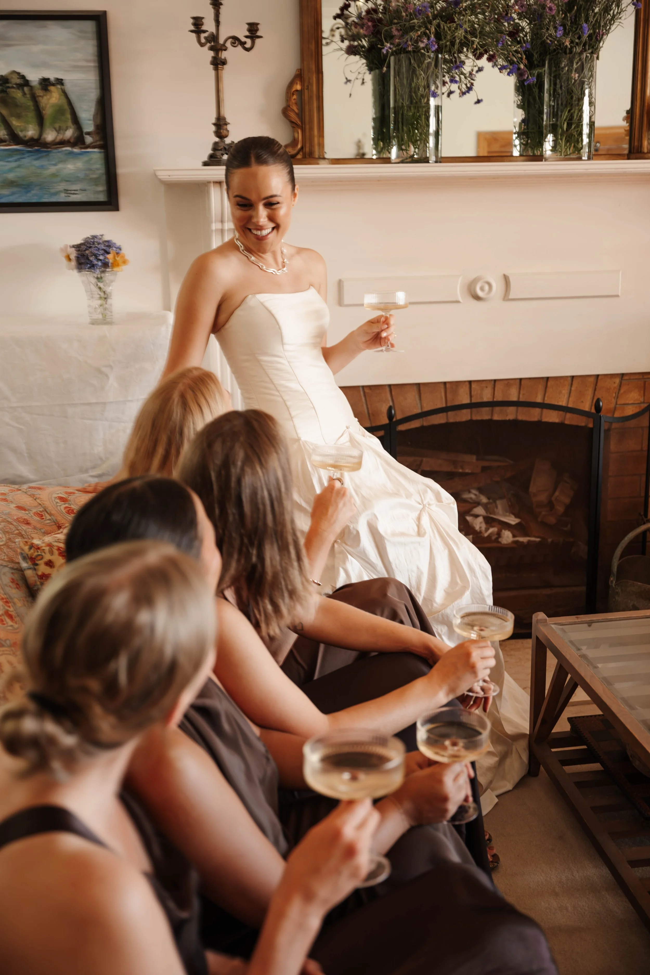 A woman in a white wedding dress holding a champagne coupe and smiling at a group of women sitting on a sofa, also holding champagne coupes, in a cozy living room with a fireplace, a large mirror, a painting, and floral arrangements.