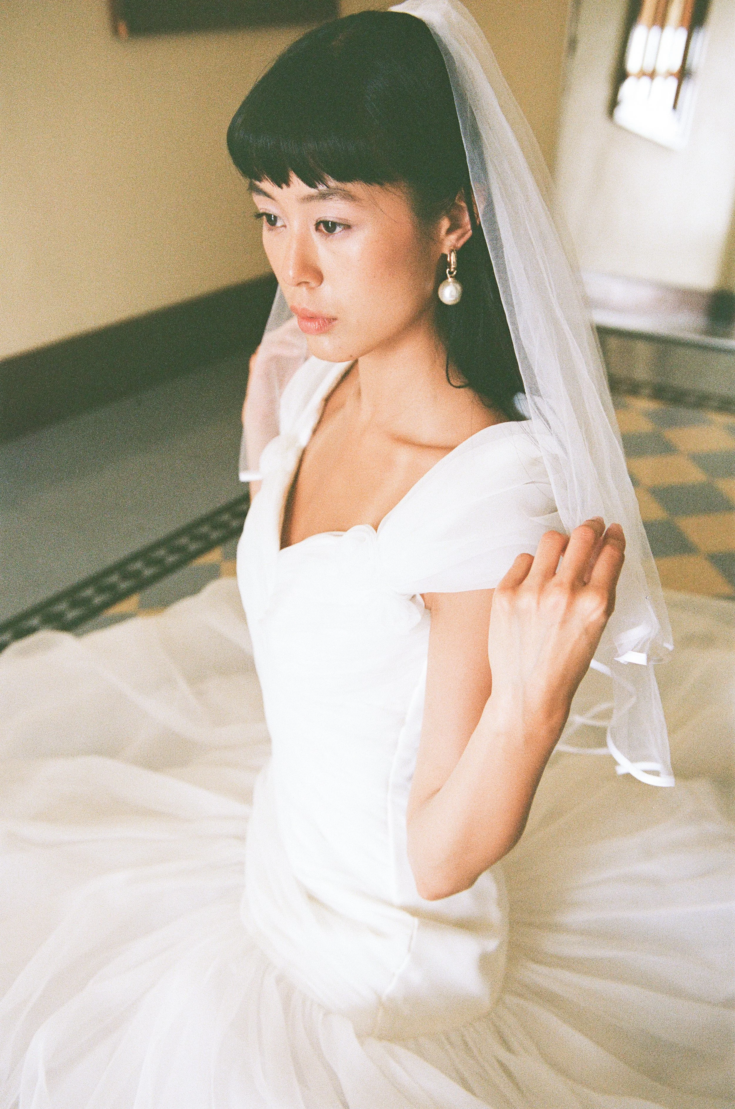 A woman dressed in a white gown with a veil, sitting on a bed, with her hand touching the veil, looking down with a contemplative expression.