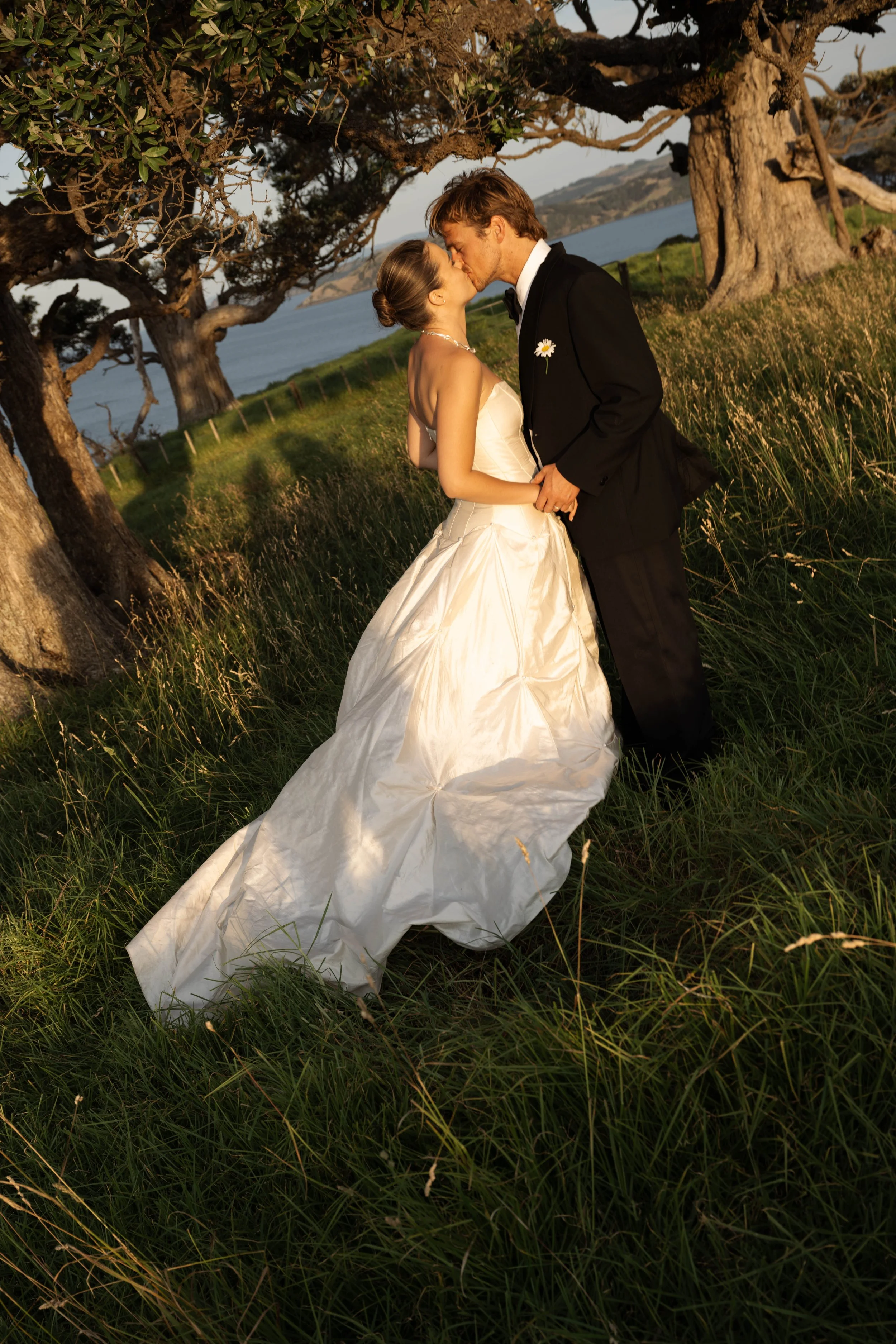 A bride and groom kissing outdoors during sunset, standing on grass near trees with a view of water and hills in the background.