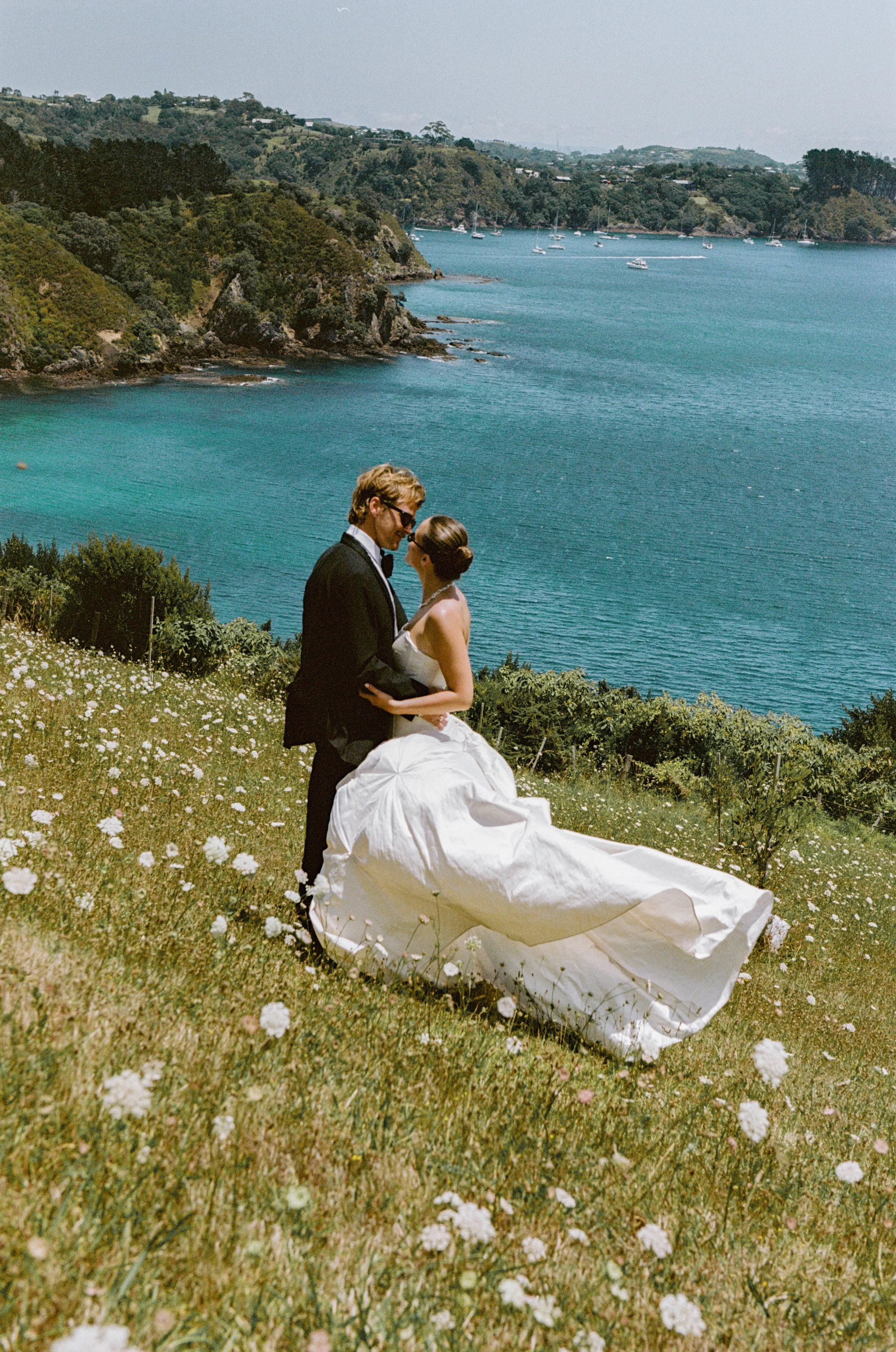 A bride and groom in wedding attire standing on a grassy hill with wildflowers, overlooking a bay with boats and green hills in the background.