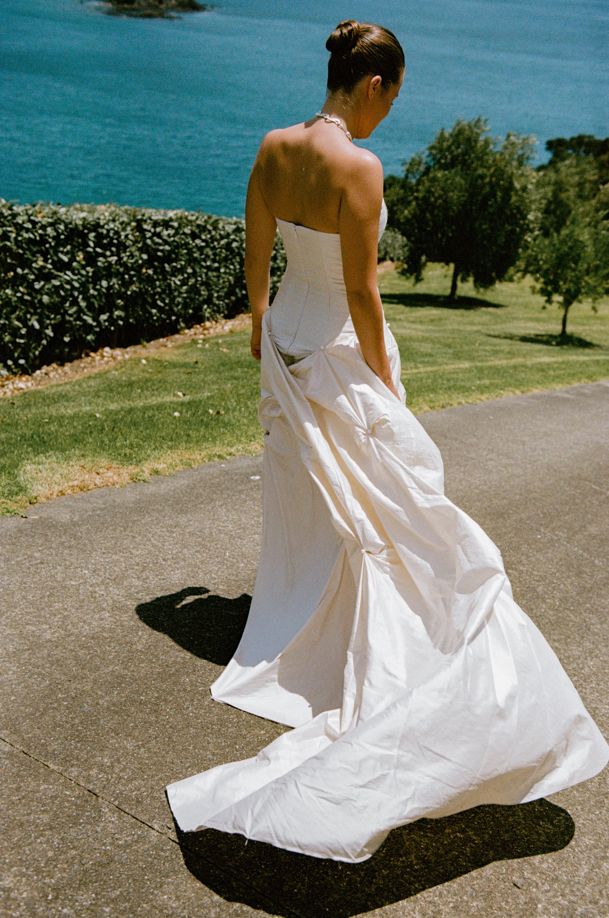 A woman in a white strapless wedding dress standing outdoors on a paved path with a view of water and trees in the background.