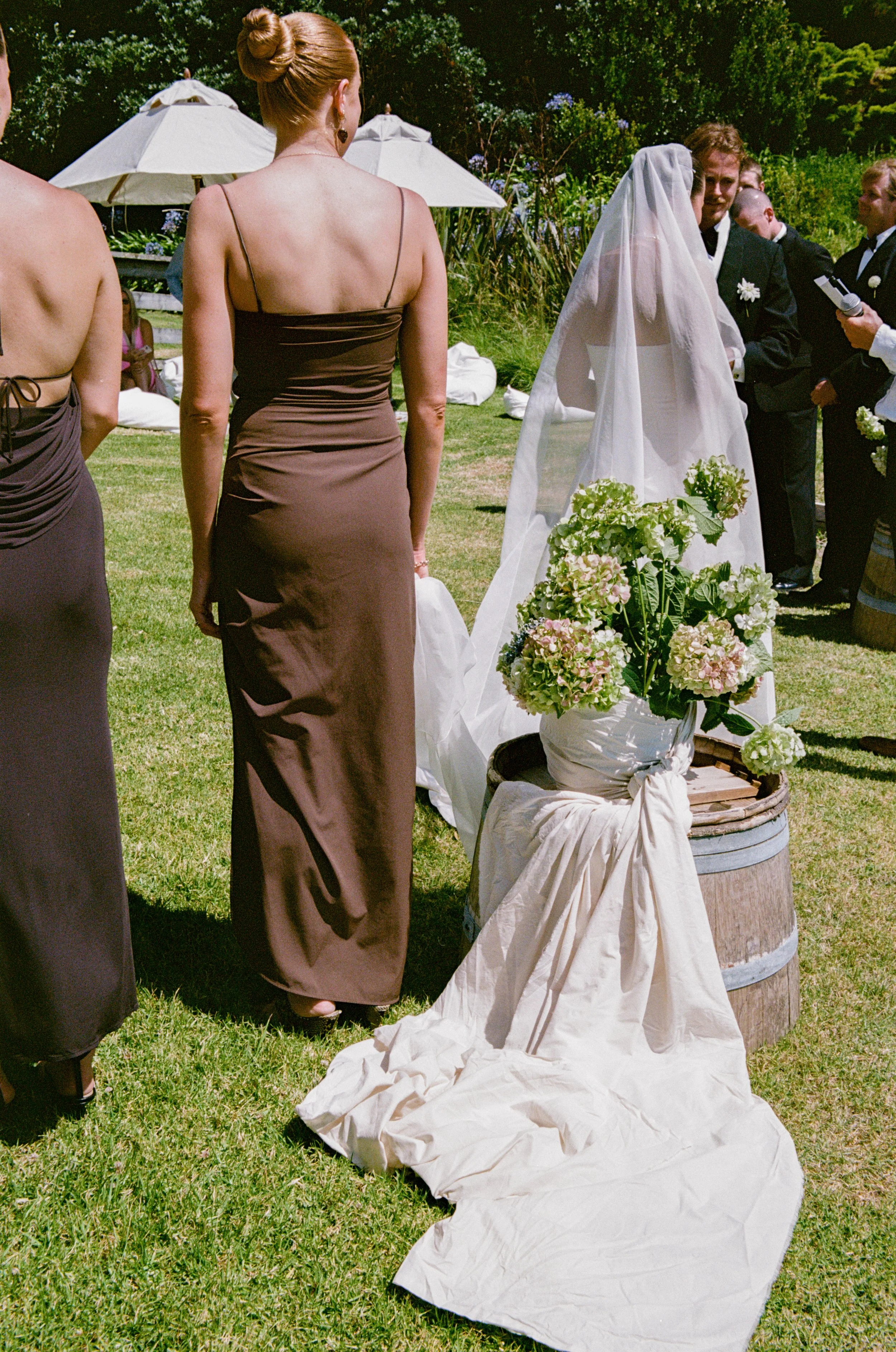 A bride in a white gown and veil standing outdoors during a wedding ceremony, surrounded by bridesmaids and groomsmen, with floral arrangements and umbrellas visible in the background.