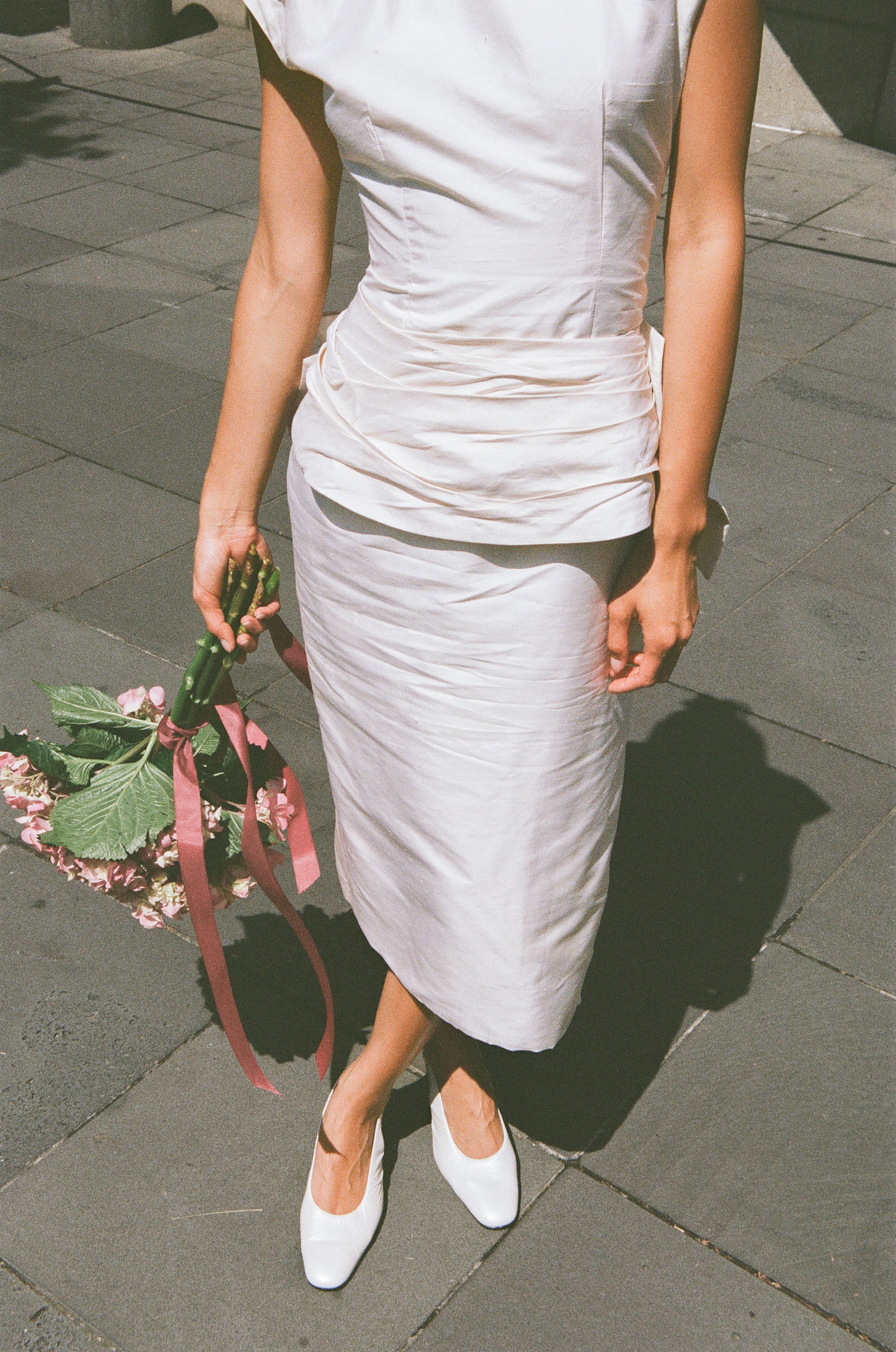A woman dressed in a white satin dress and white shoes is holding a bouquet of pink and green flowers with a pink ribbon, standing on a paved sidewalk.