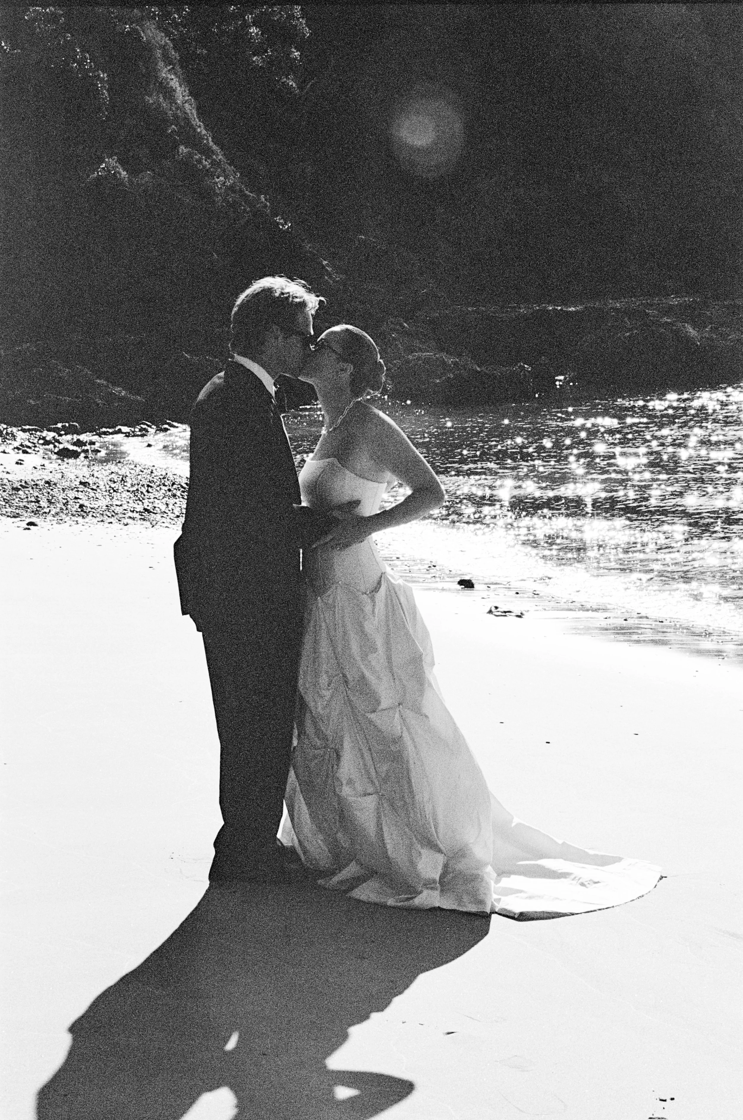 A black and white photo of a couple in wedding attire kissing on a beach at night, with the ocean and rocky cliffs in the background.