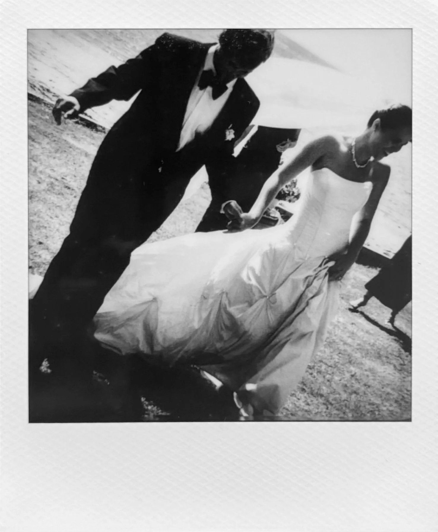 Black and white photo of a man and a woman in formal attire standing outdoors on a beach, holding hands and leaning over a large cloth or sheet.