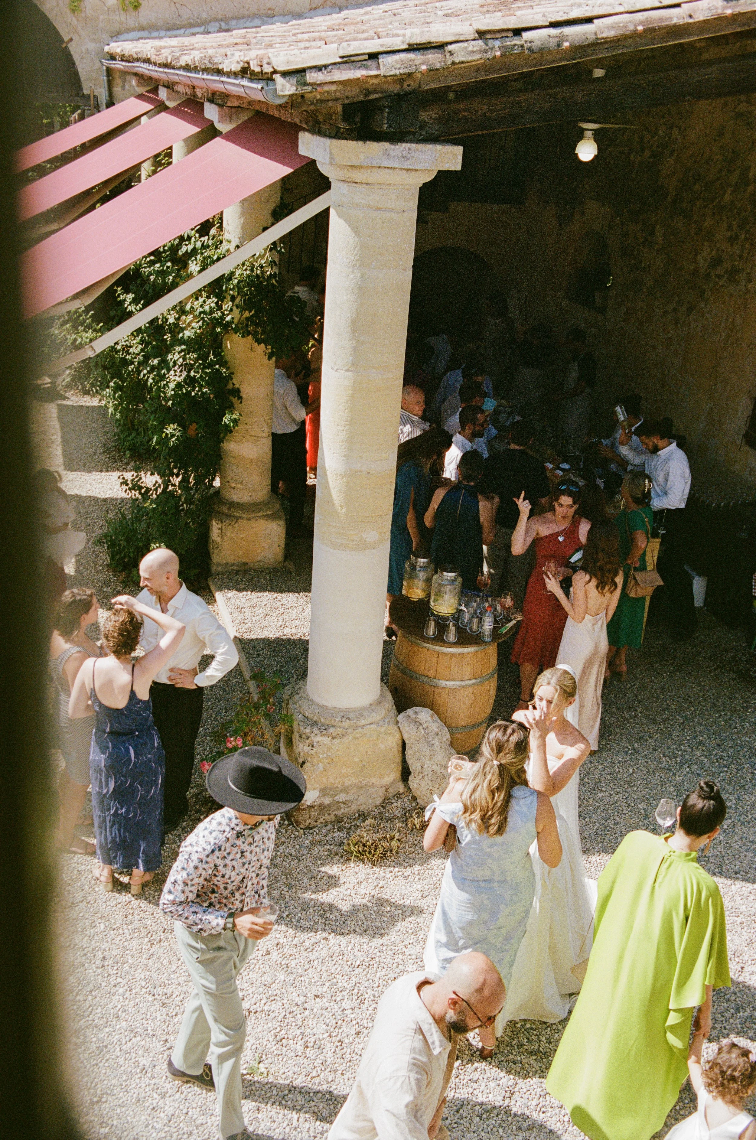 People gathered in an outdoor courtyard, socializing near a bar area with drinks and tables under a stone archway with picnic tables, awnings, and greenery.