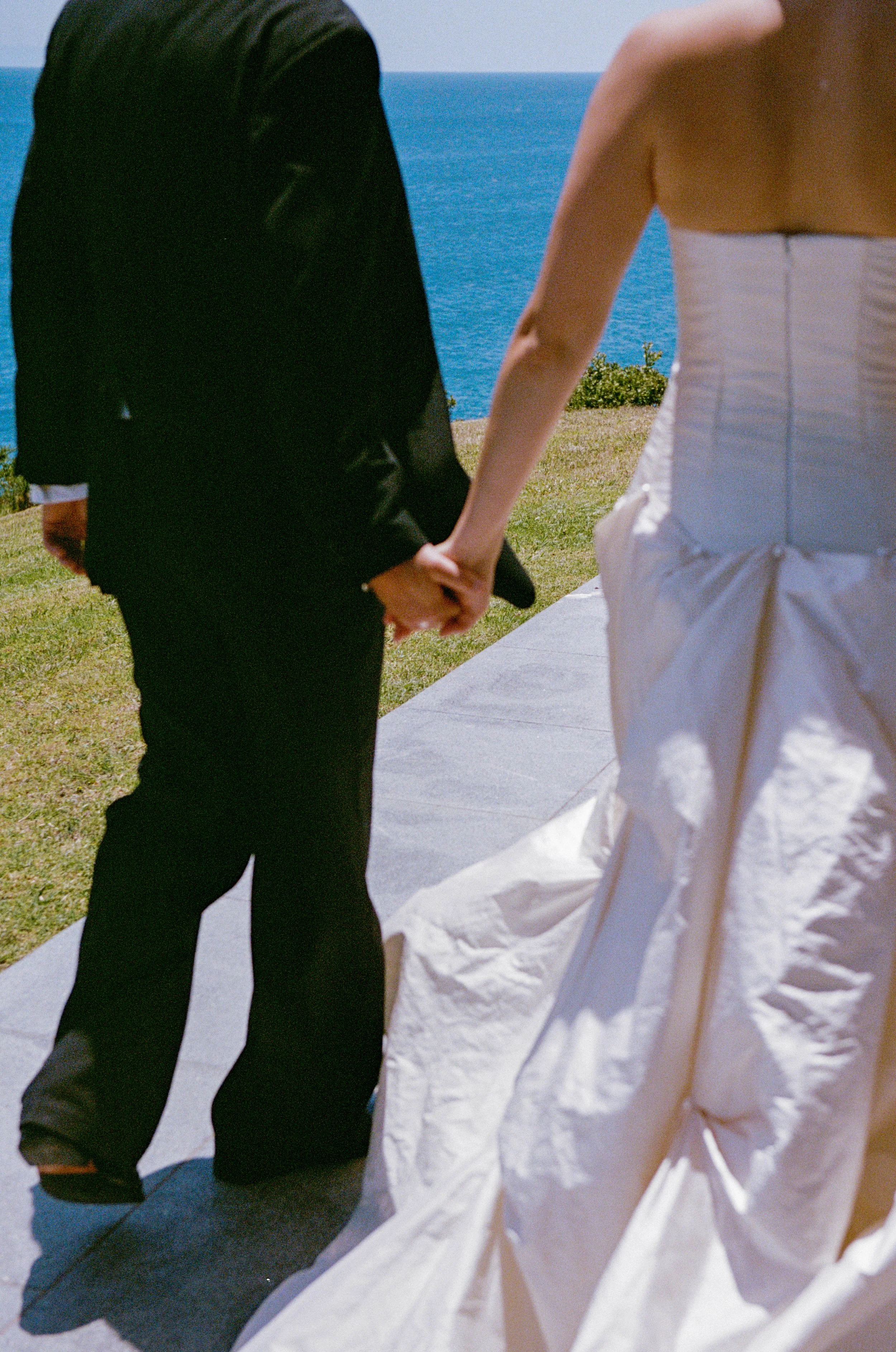 A couple holding hands at an outdoor wedding ceremony near the ocean, with one person in a black suit and the other in a white wedding dress.