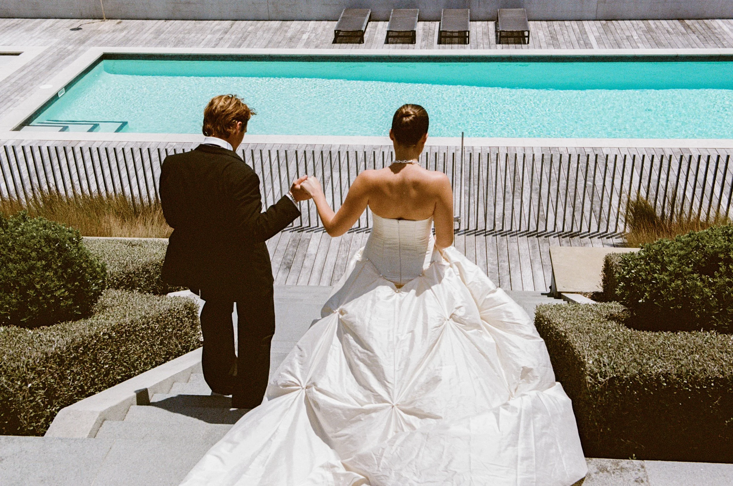A wedding bride in a white wedding dress holding hands with the groom, who is dressed in a black suit, as they stand on stairs near a swimming pool with modern lounge chairs in the background.
