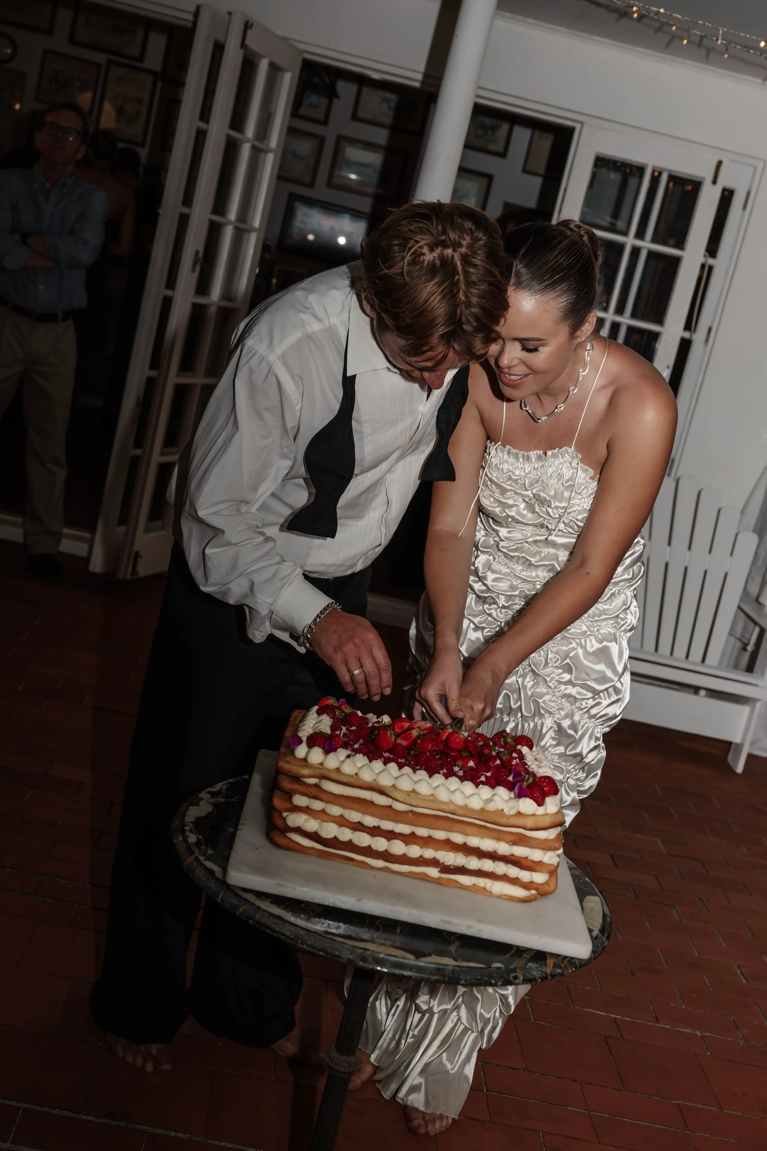A woman in a satin dress and a man in a white shirt and black pants cutting a wedding cake decorated with strawberries, raspberries, and purple flowers at a celebration event.