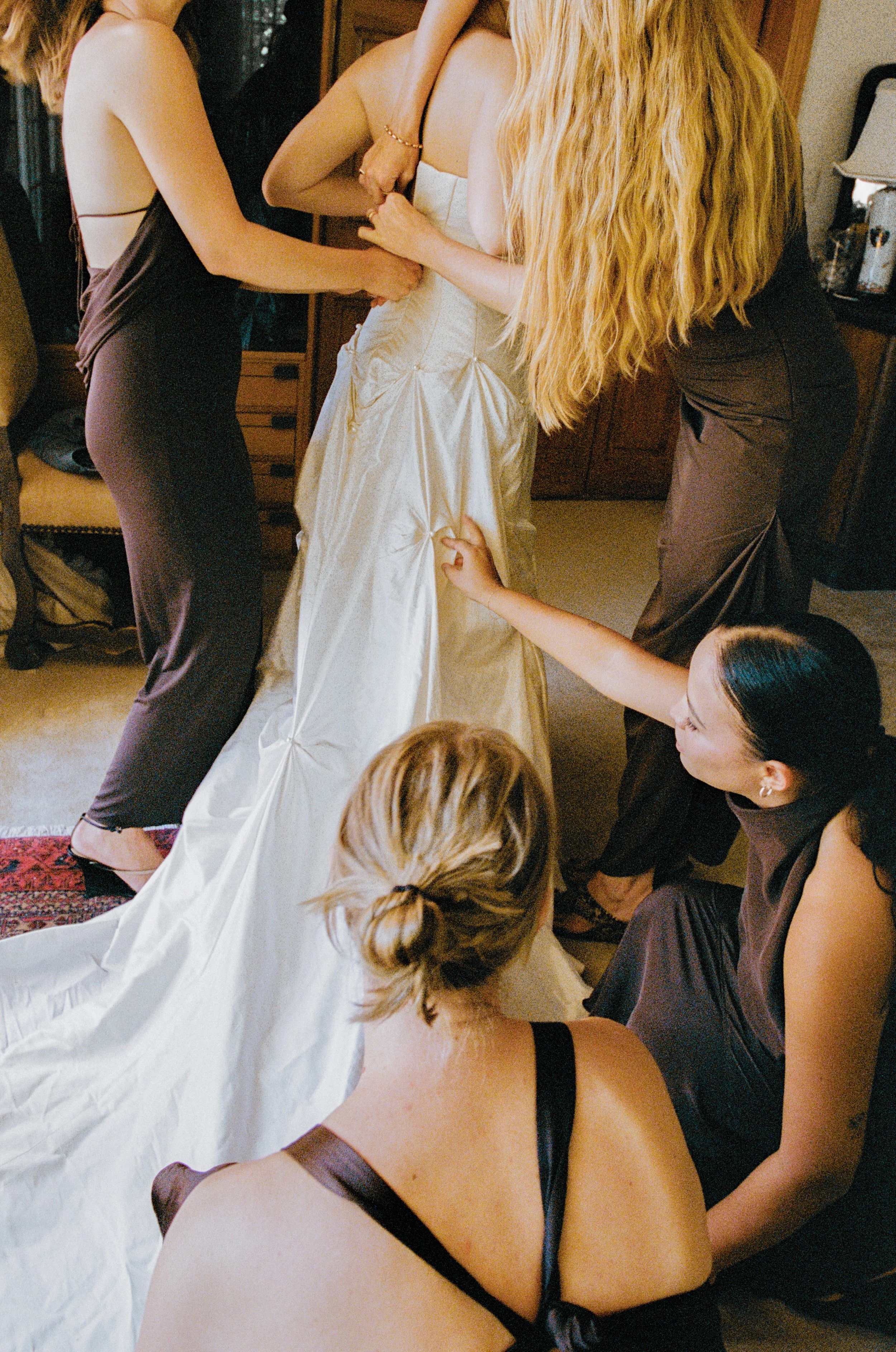 A bride in a wedding dress is being assisted by five women to prepare for her wedding.