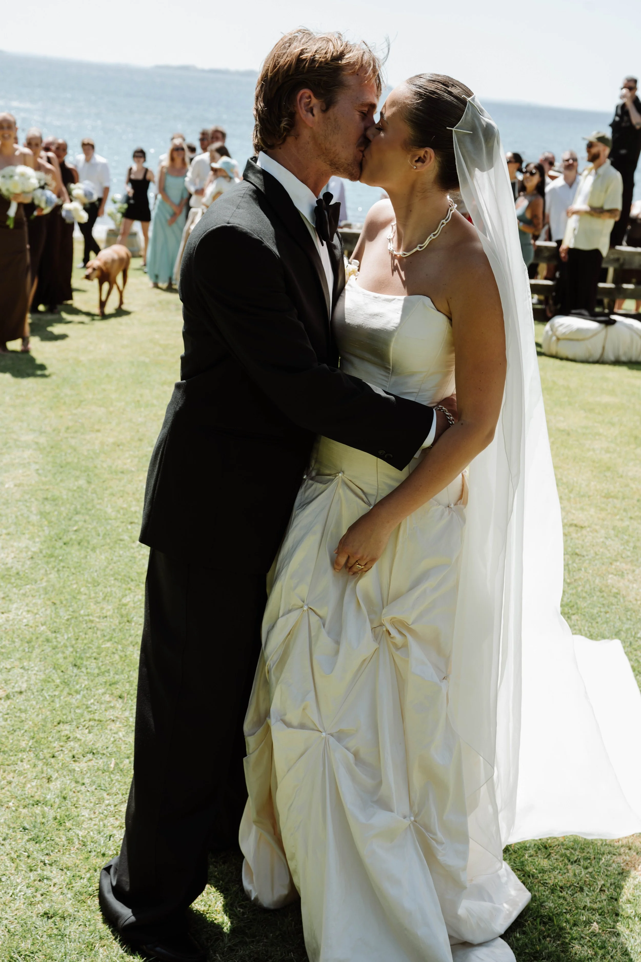 A bride and groom kiss during their outdoor wedding ceremony by the water, with guests and a dog in the background.