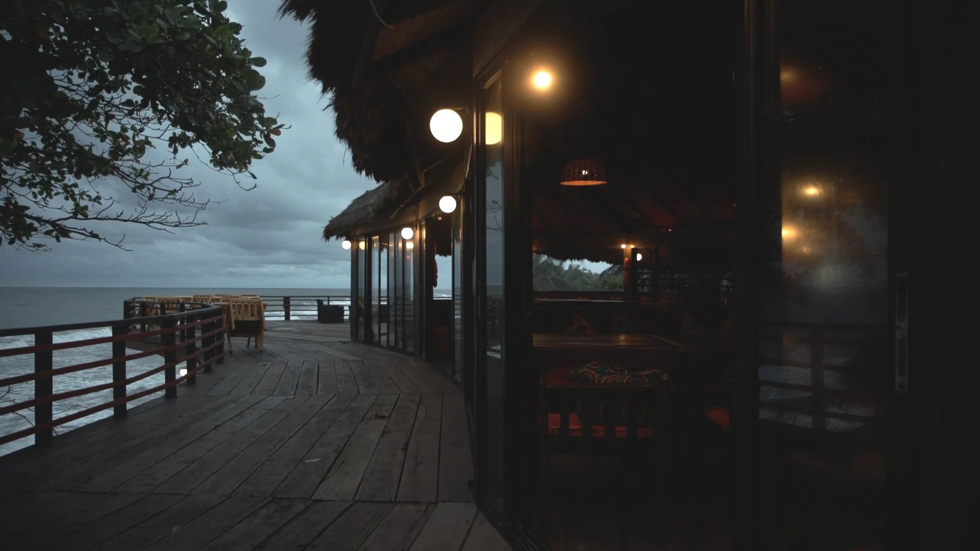 Oceanfront deck at dusk with a hut featuring large glass windows and warm interior lighting, surrounded by trees and cloudy sky