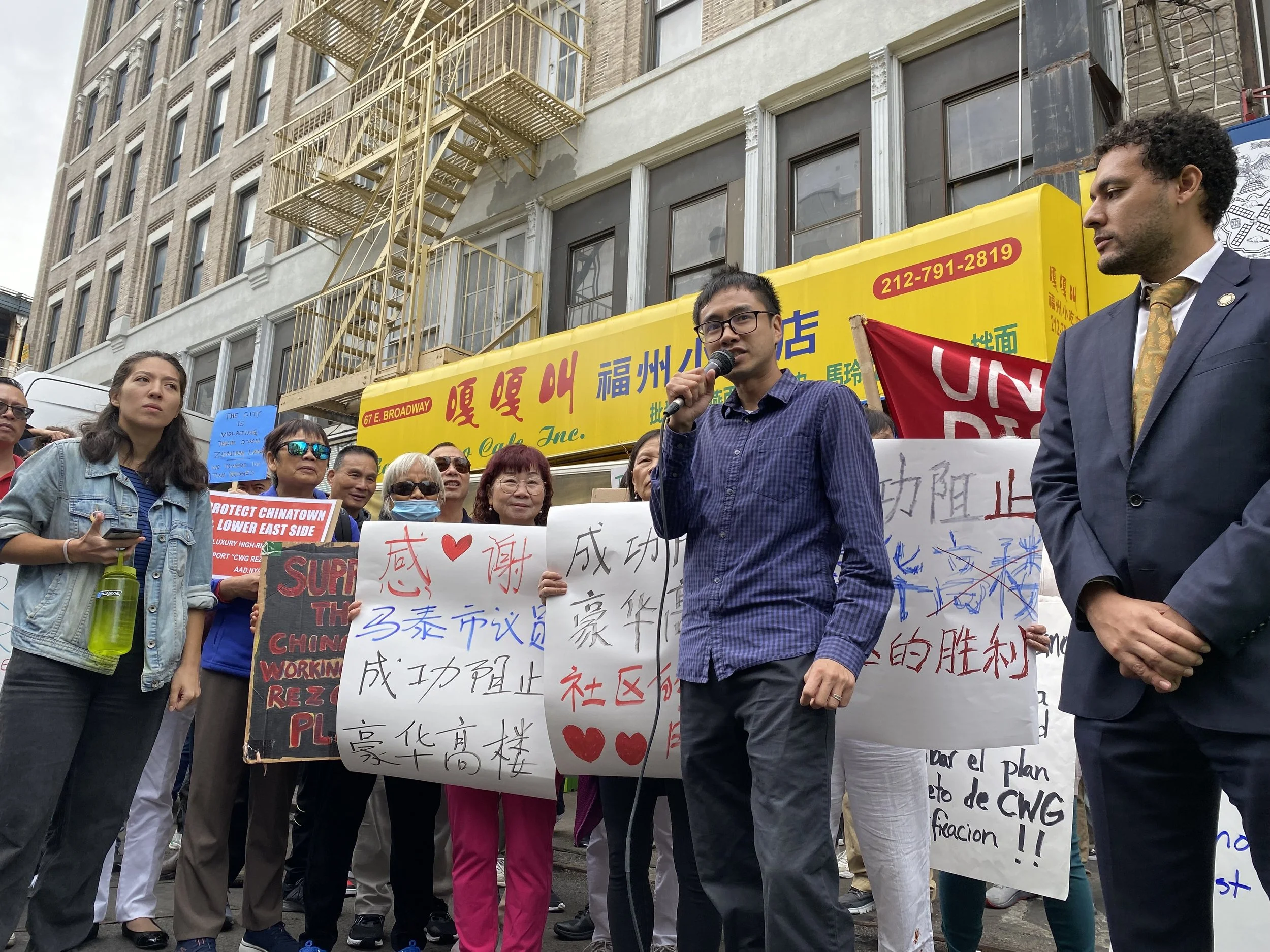 A group of diverse people gathered outdoors on city street, participating in a protest or rally with signs and banners, some holding signs in Chinese and English, including messages supporting Chinatown and against gentrification, with buildings and a yellow storefront in the background.