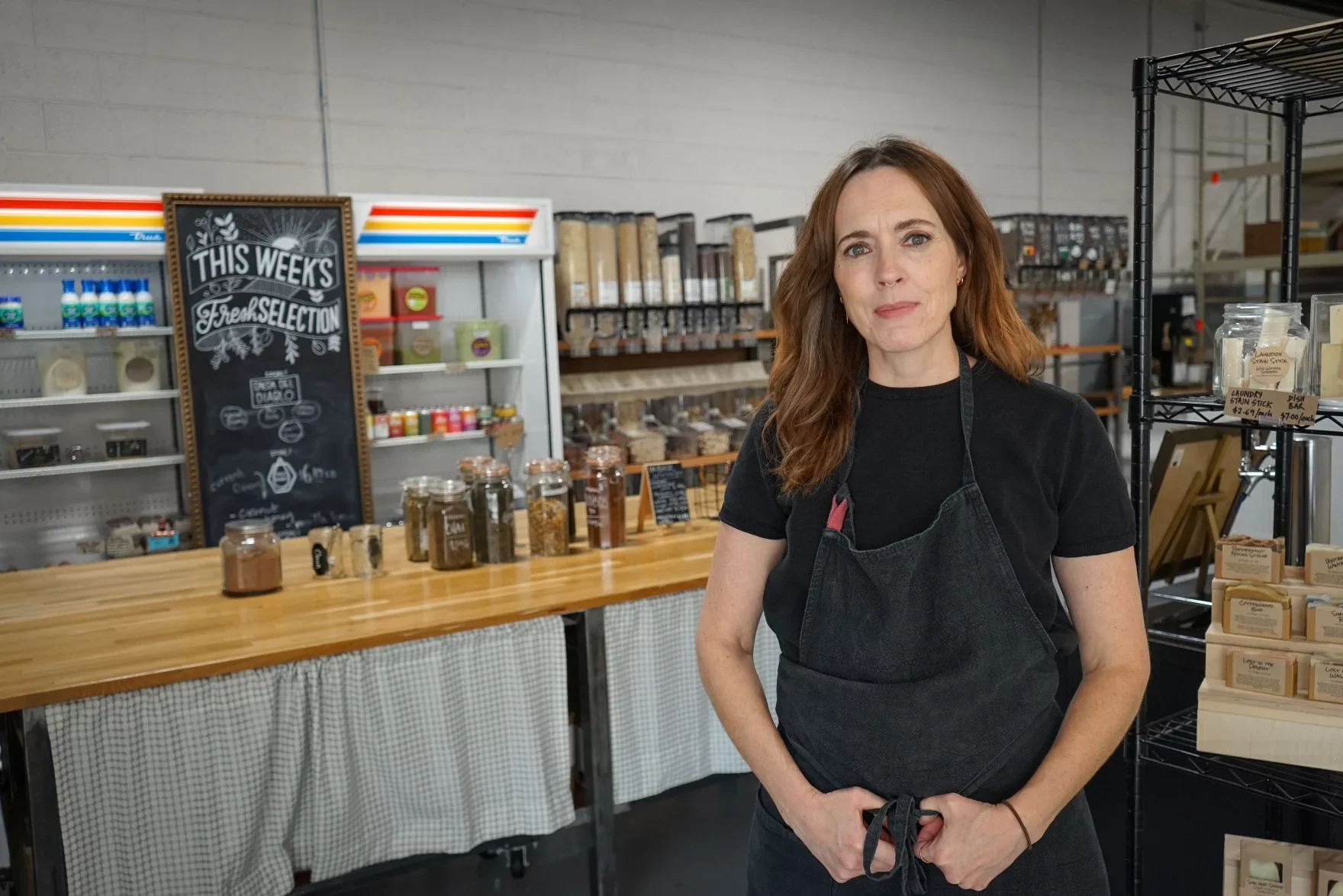 A woman with reddish-brown hair wearing a black T-shirt and a black apron standing behind a wooden counter in a grocery store, with jars of spices and a sign that reads 'This Week's Fresh Selection' in the background.
