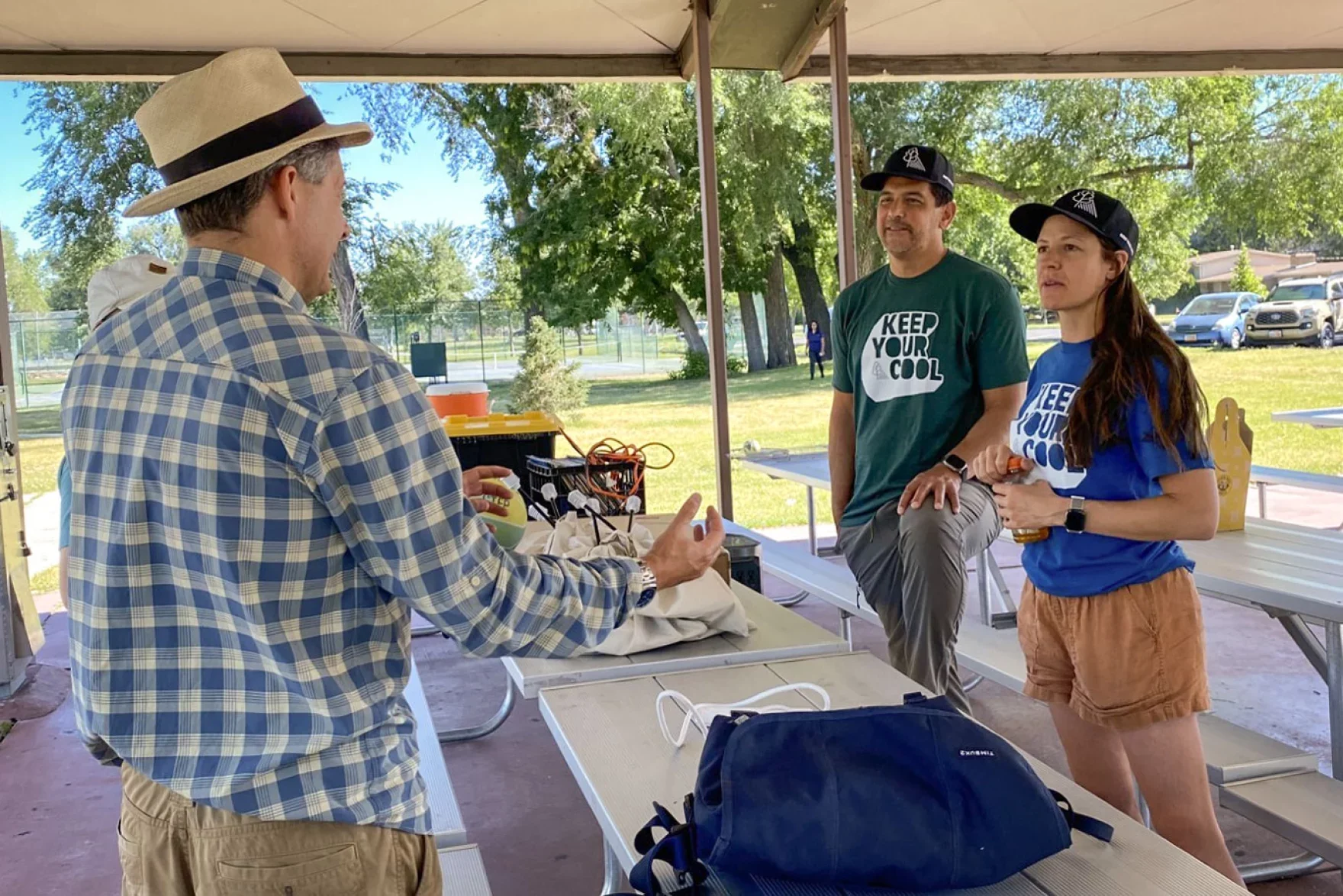 Three people engaged in a conversation outdoors under a canopy, with a picnic table and parked cars in the background. Two of them are wearing blue and green T-shirts with 'Keep Your Cool' printed on them, and the third person, facing away from the camera, is wearing a plaid shirt and a straw hat.