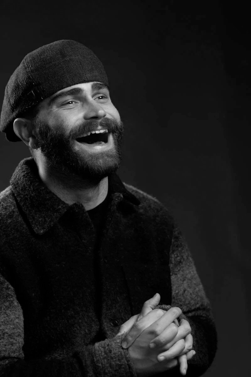 A black and white photo of a man with a beard and mustache, wearing a beanie and a textured jacket, smiling and laughing with clasped hands.