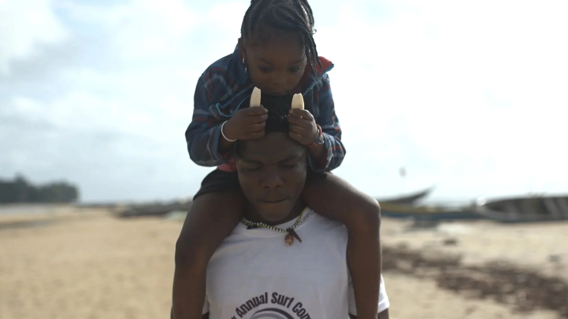 A person giving a piggyback ride to a child on a beach with boats in the background.