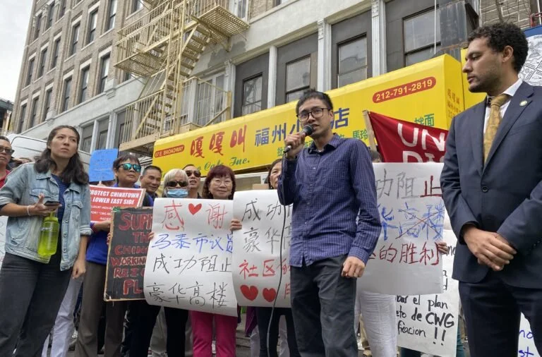 Group of people holding signs and listening to a man speaking into a microphone on a city street.