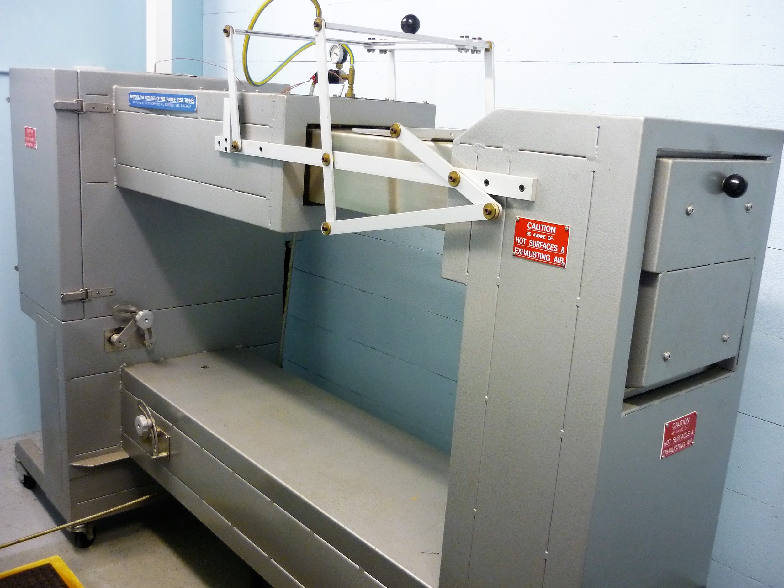 Laboratory equipment for testing, mounted on a metallic frame with warning labels indicating hot surfaces and exhausting air.