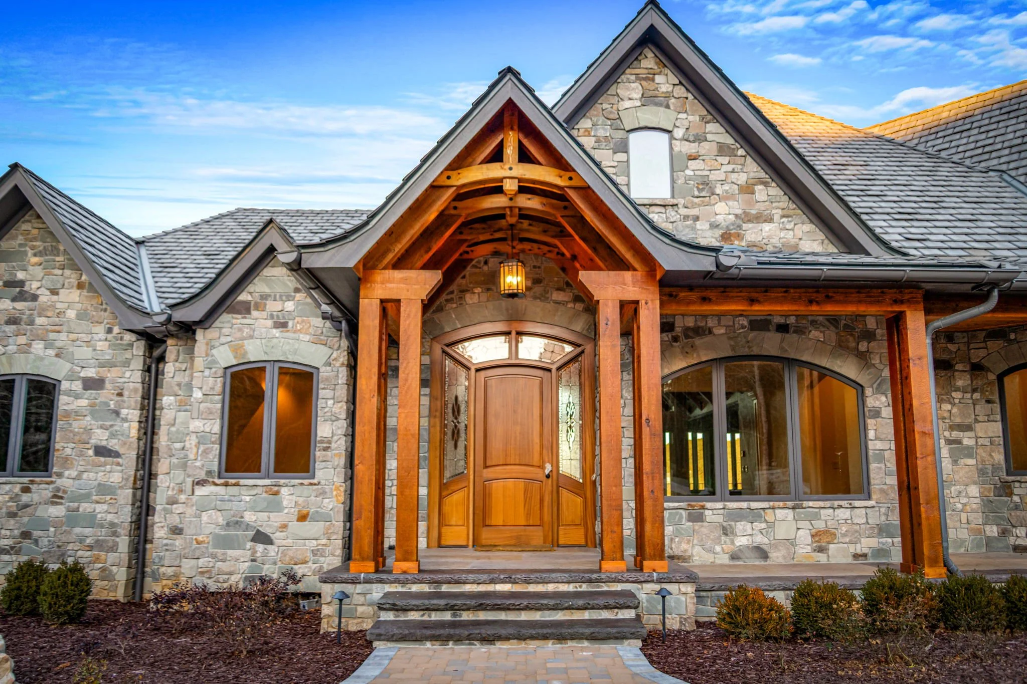 Front view of a house with stone walls, a wooden front door, and a porch with wooden beams, under a blue sky with some clouds.
