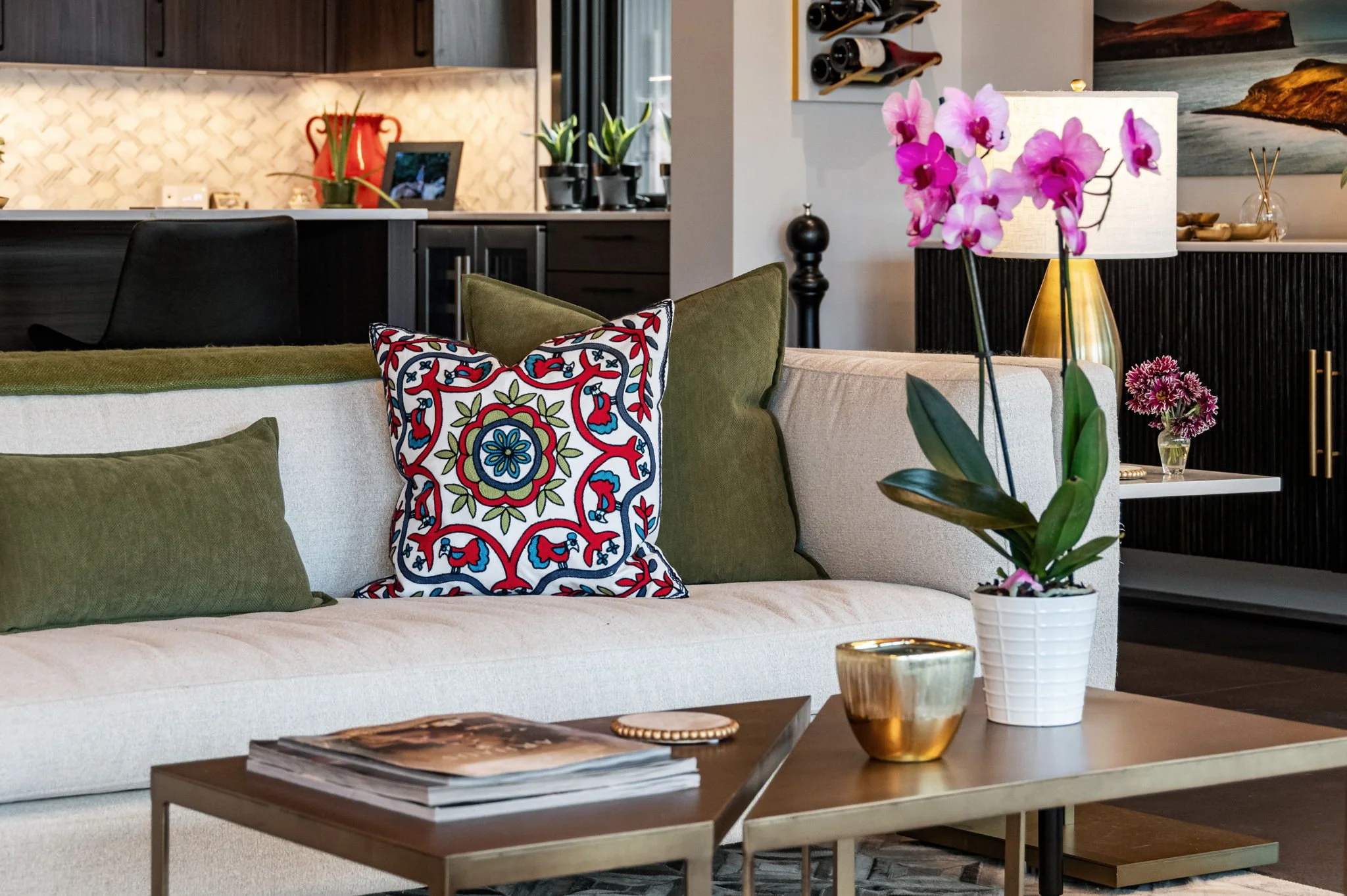 A modern living room with a beige sofa decorated with green and patterned pillows, a wooden coffee table with magazines, a potted orchid, a gold-colored candle holder, and a lamp with a white shade. In the background, there is a kitchen area with black cabinets, framed artwork, plants, and wine bottles.