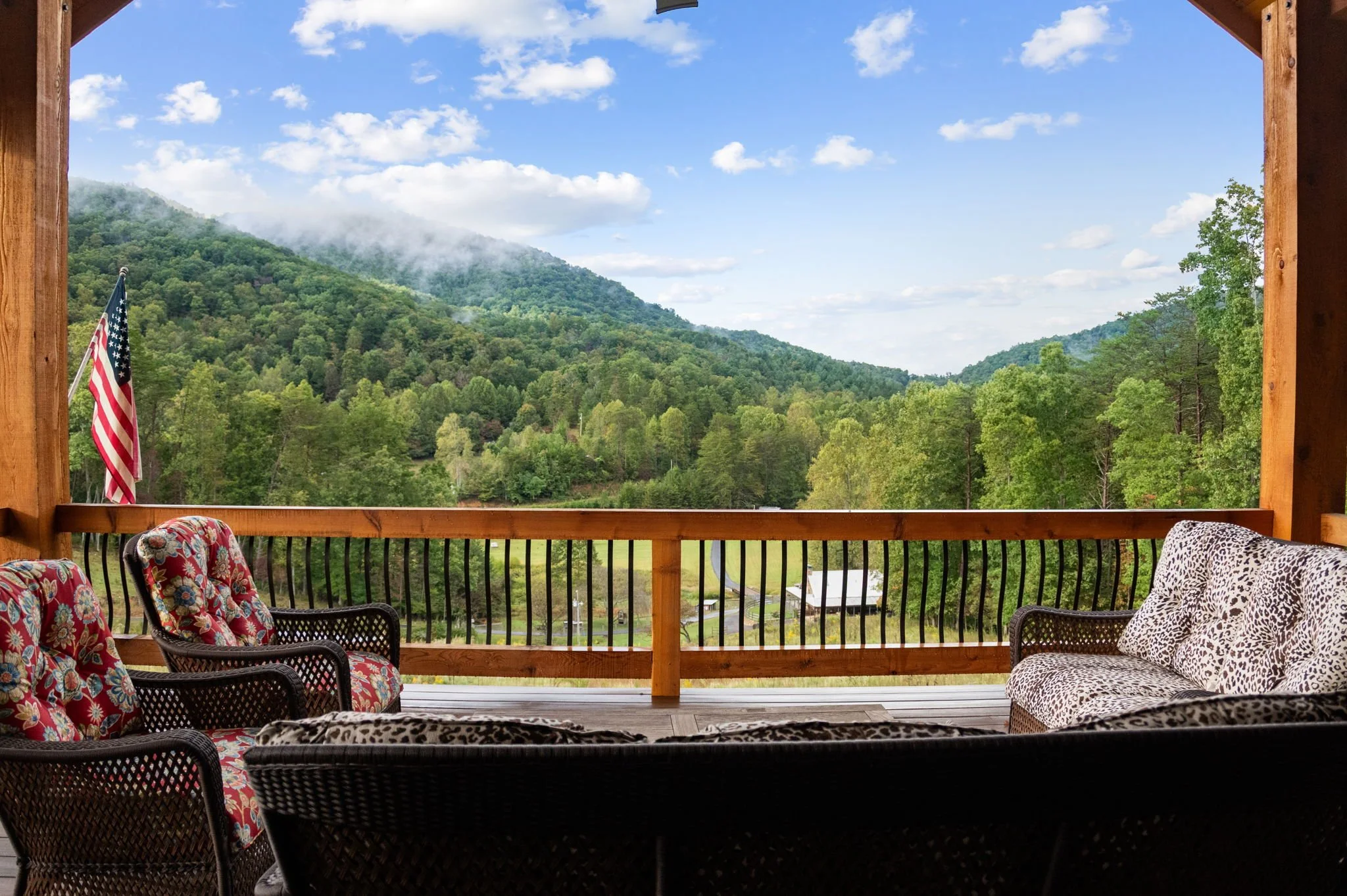 View of a lush, green mountain landscape from a wooden porch with outdoor furniture, including cushioned chairs and a leopard print sofa, and an American flag.