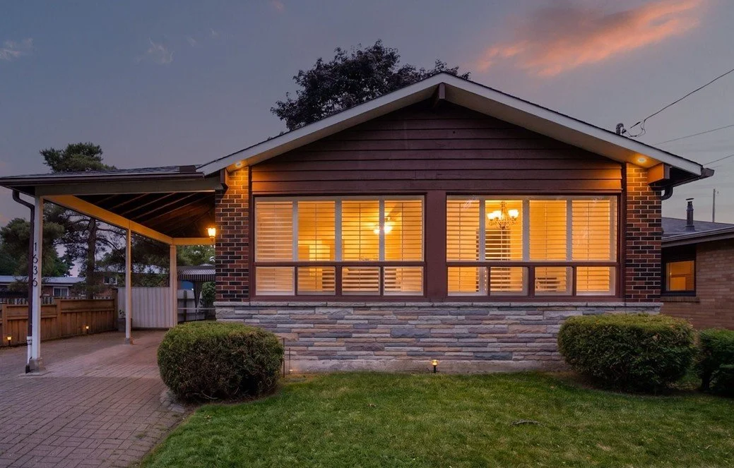 Front view of a house during sunset with illuminated windows, a brick and stone exterior, a covered driveway on the left, and landscaped bushes in front.