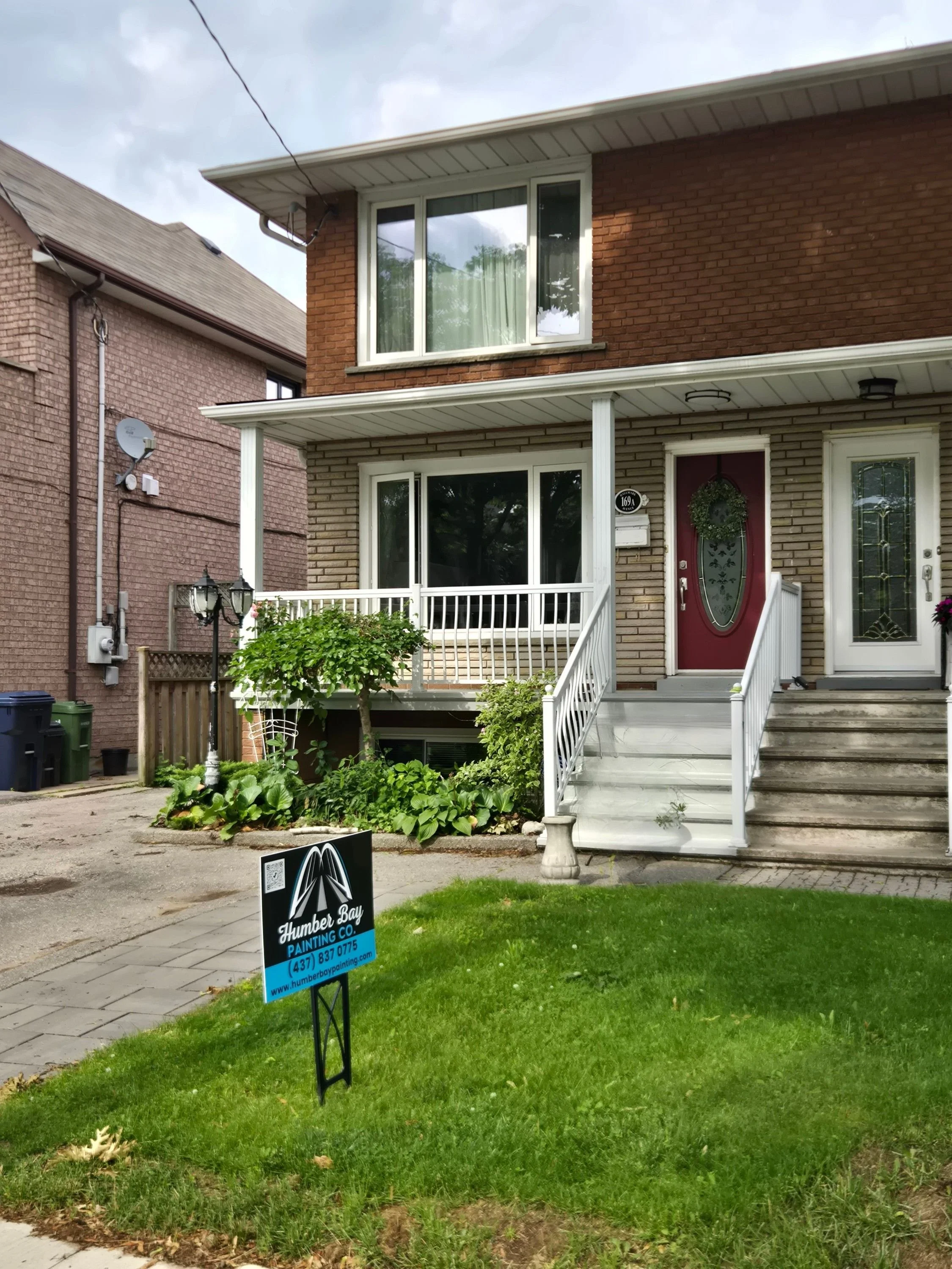 A two-story brick house with a front porch and stairs leading up to the front door, surrounded by a neatly maintained lawn and some plants. A sign on the lawn indicates the house is being painted by Humber Bay Painting Co., and there is a neighboring