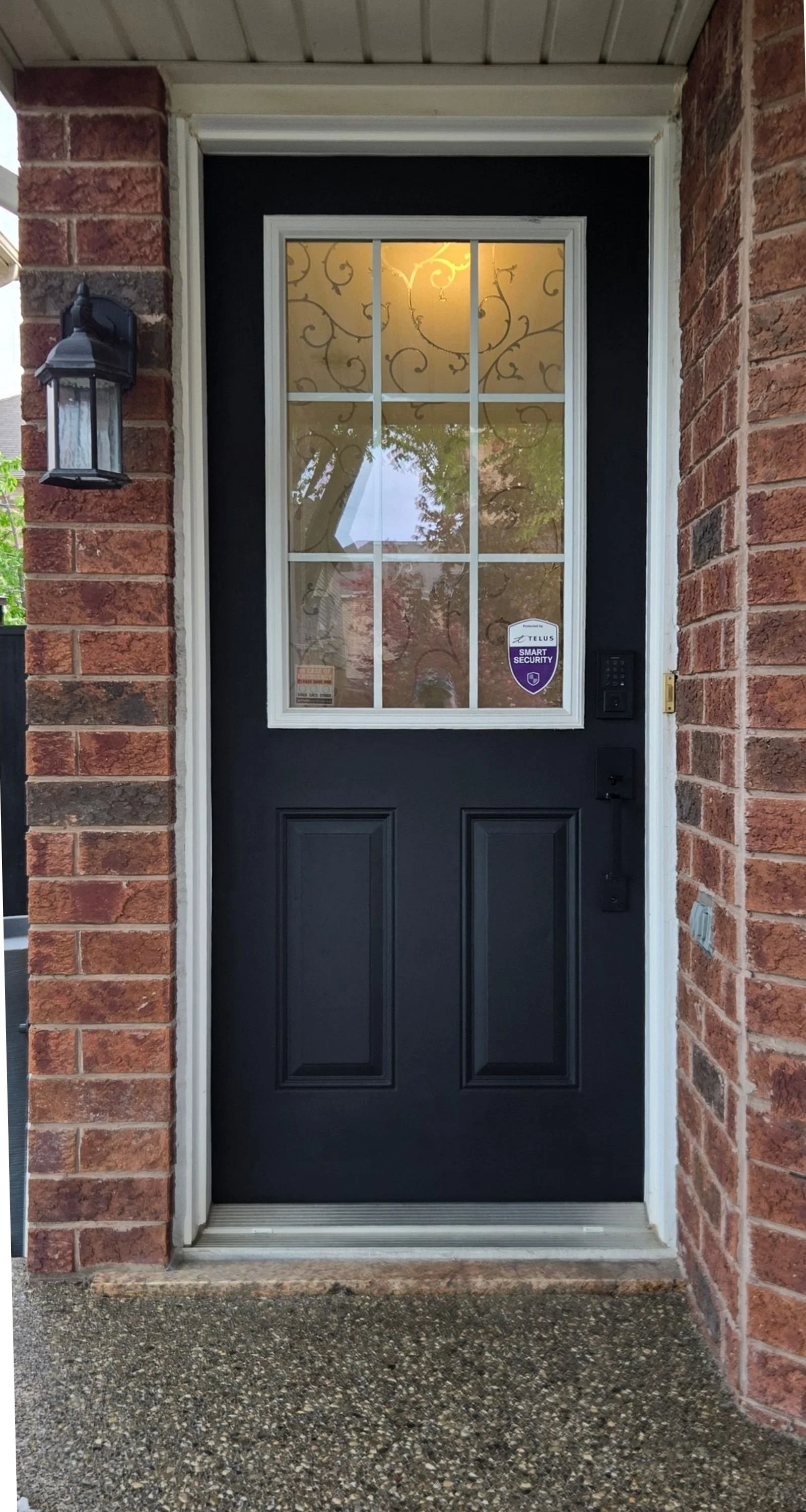 Black front door with a window divided into six panes, set in a brick wall and topped with forest green siding. A black security sticker is on the window, and a black doorbell with a camera is installed on the right side. An outdoor lantern light fix