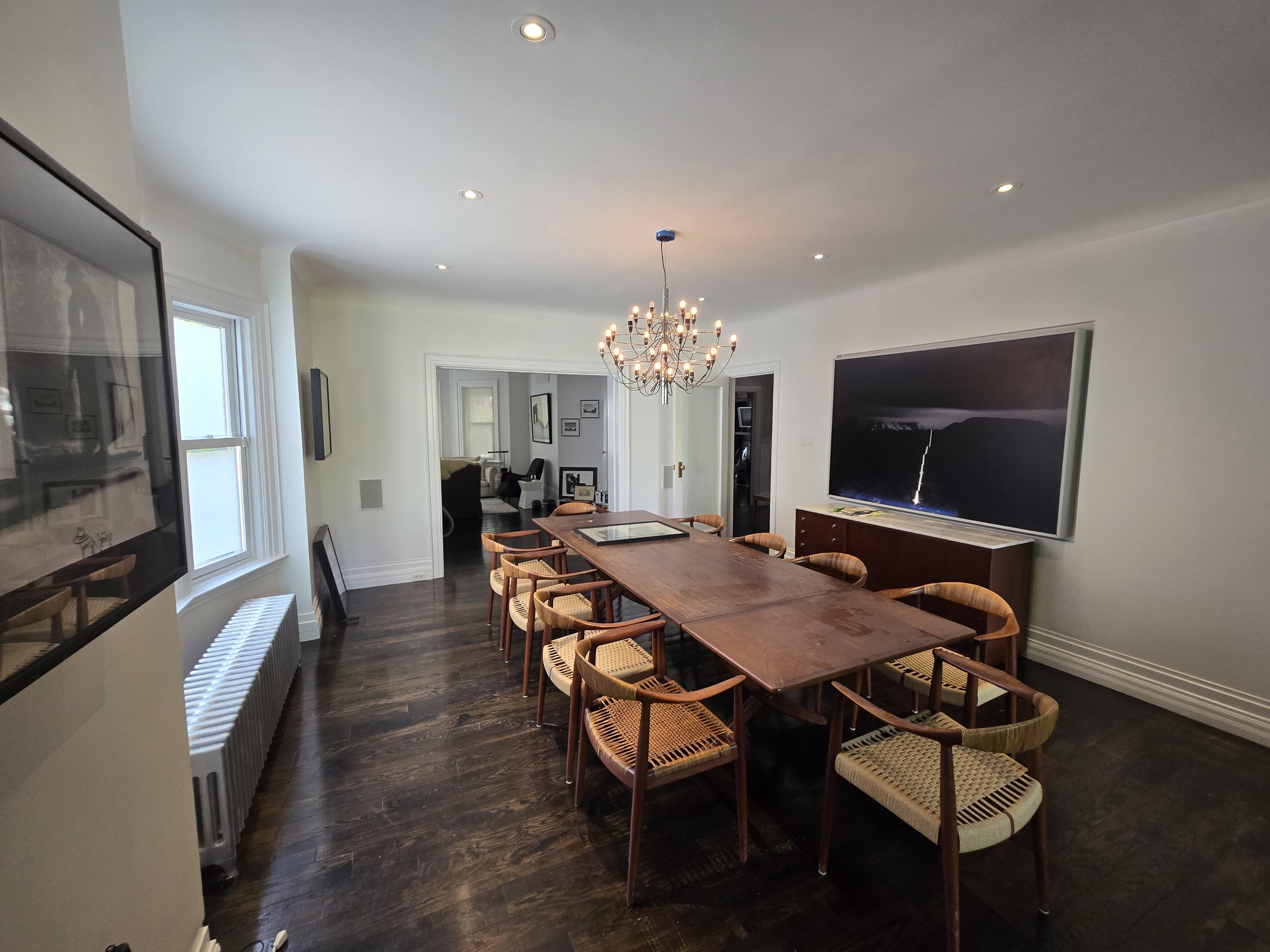 A dining room with a wooden table surrounded by eight chairs with woven seats. There is a chandelier hanging above the table, and a large flat-screen TV mounted on the wall. The room has white walls, dark wood flooring, and windows letting in natural