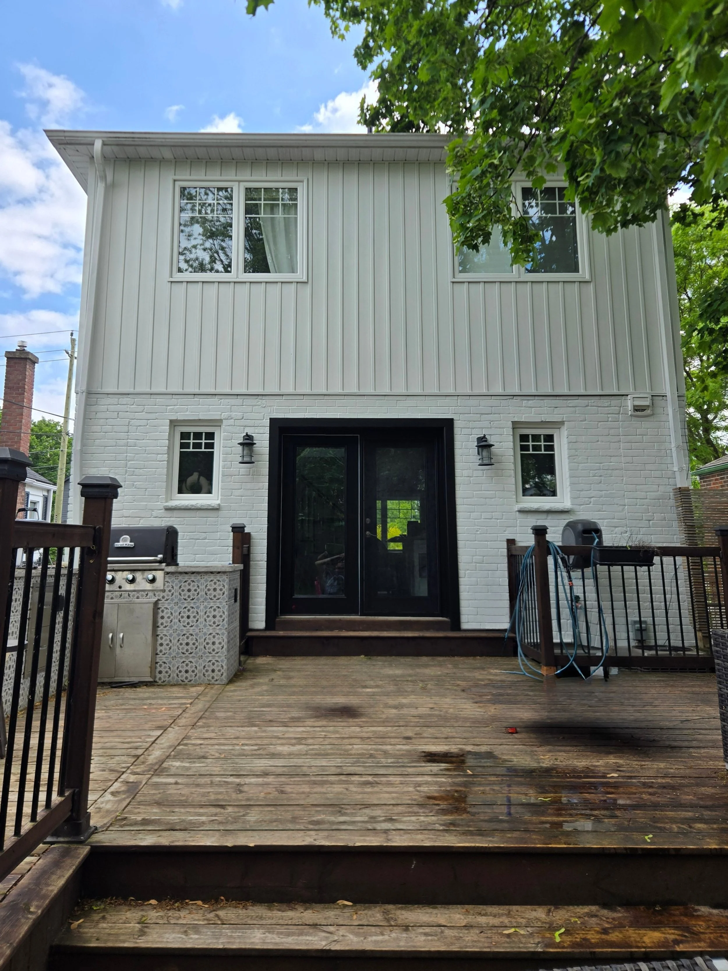 Backyard deck with wooden floorboards, a black screen door leading into the house, small side windows, outdoor wall-mounted lights, a grill, garden hose, and fencing, with trees and blue sky in the background.