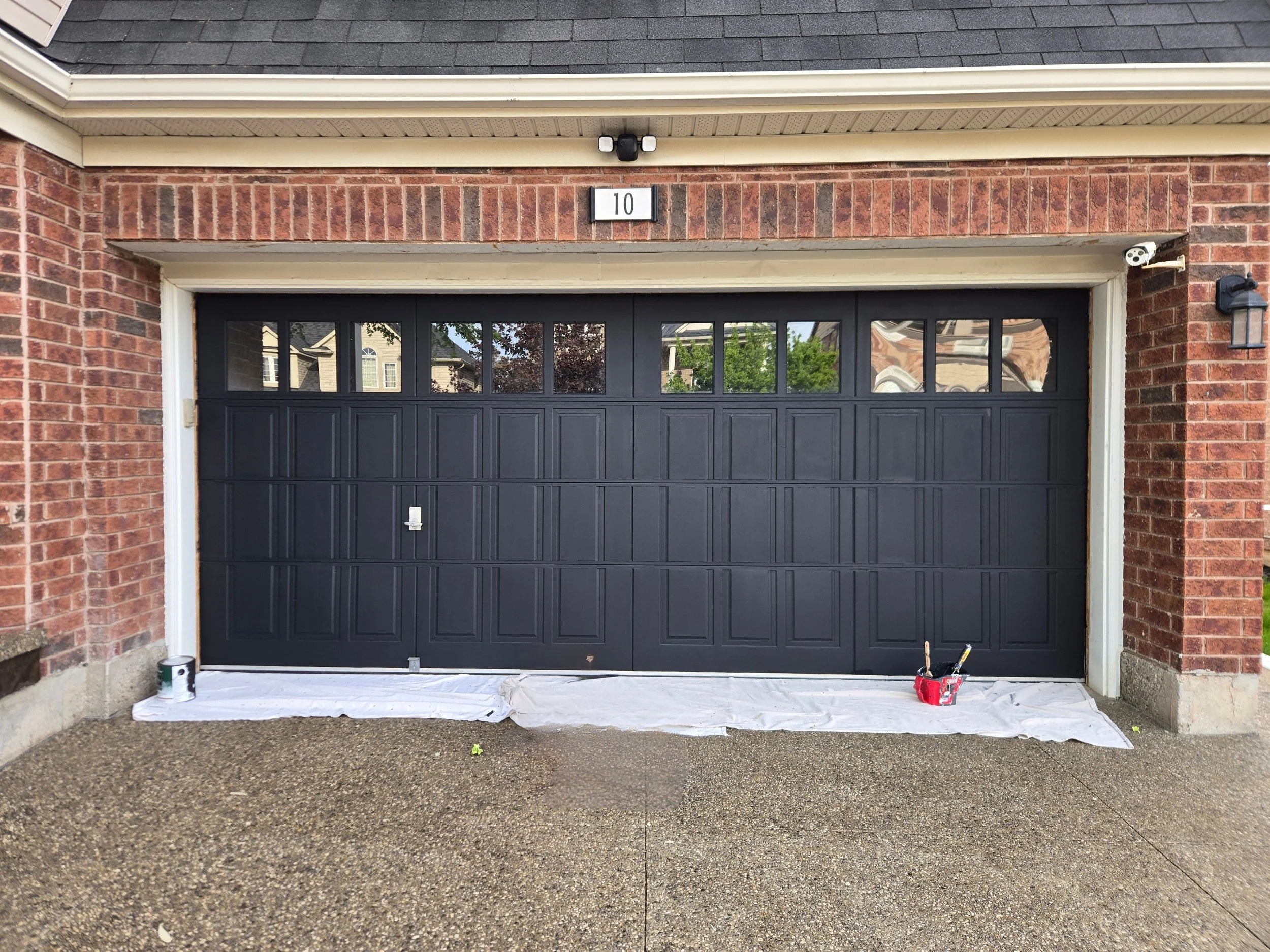 The image shows a black garage door with four small windows near the top, set in a brick house. Above the garage, there is a brick wall with house number 10 and an outdoor camera on each side. The ground in front of the garage is covered with a white