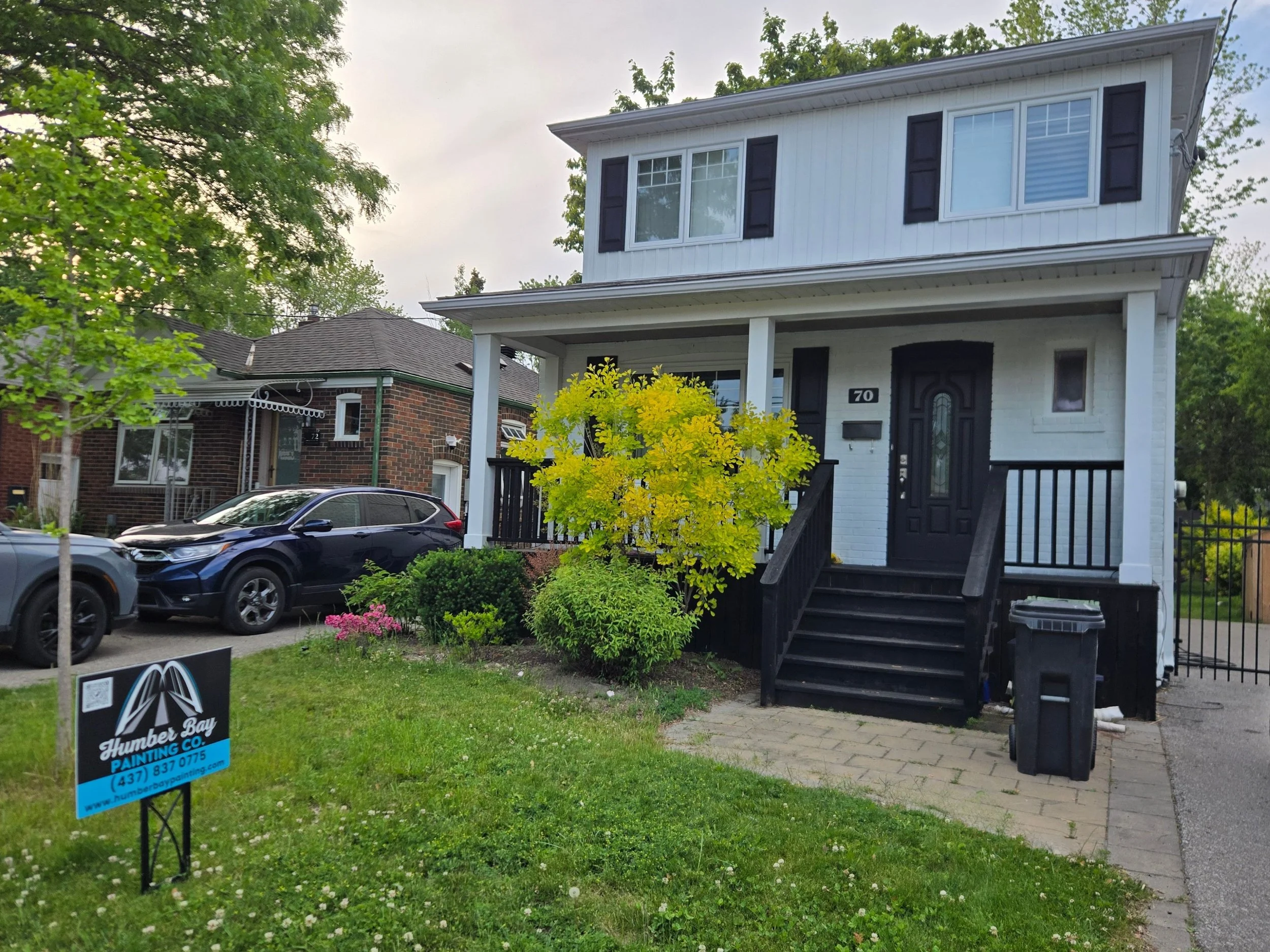 Front view of a white two-story house with black shutters and a dark front door. A small porch with stairs leads to the door. There are trees with green leaves and a small garden with yellow and pink flowers in front. A black car is parked in the dri