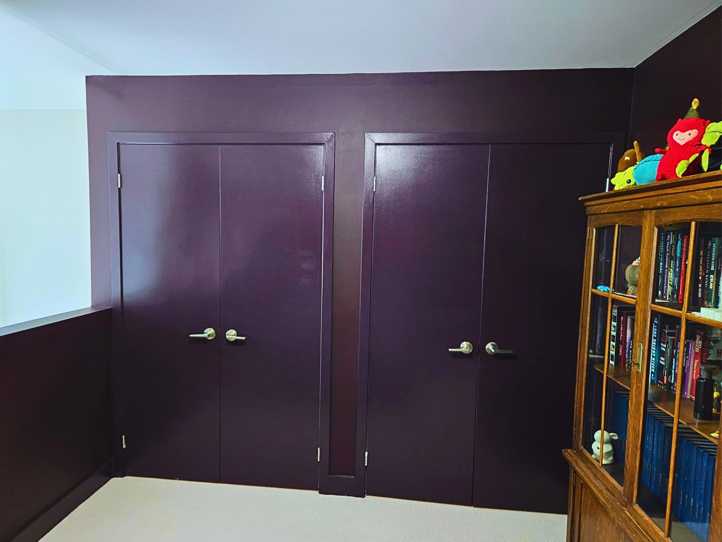 A room with dark purple closet doors, a black wall, a light ceiling, a white wall on the left, a beige carpet, and a wooden bookshelf on the right filled with books and toys.