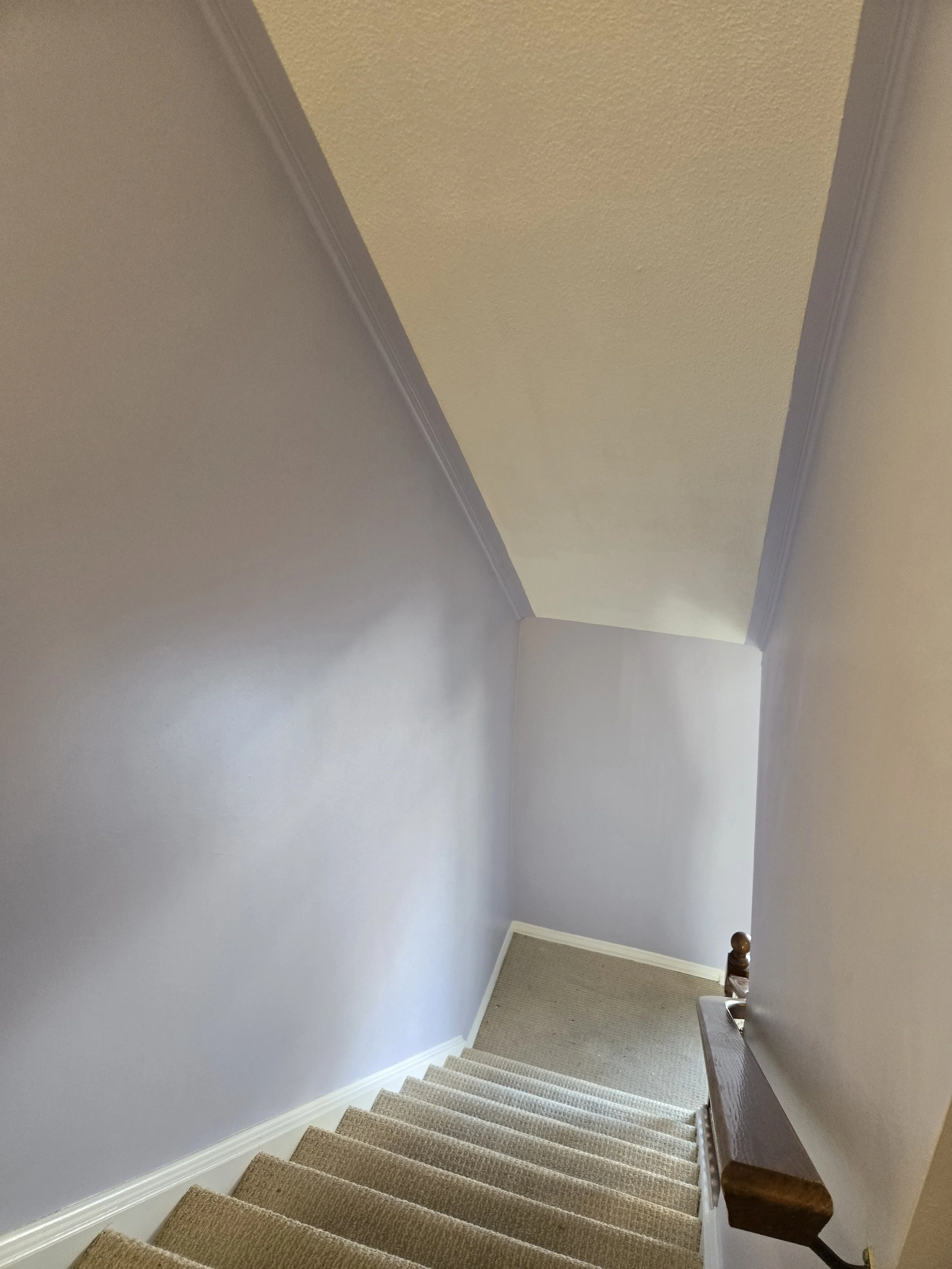 A staircase with beige carpet leading down to a landing with a small table and a brown railing on the right side, with a yellow ceiling and light gray walls.