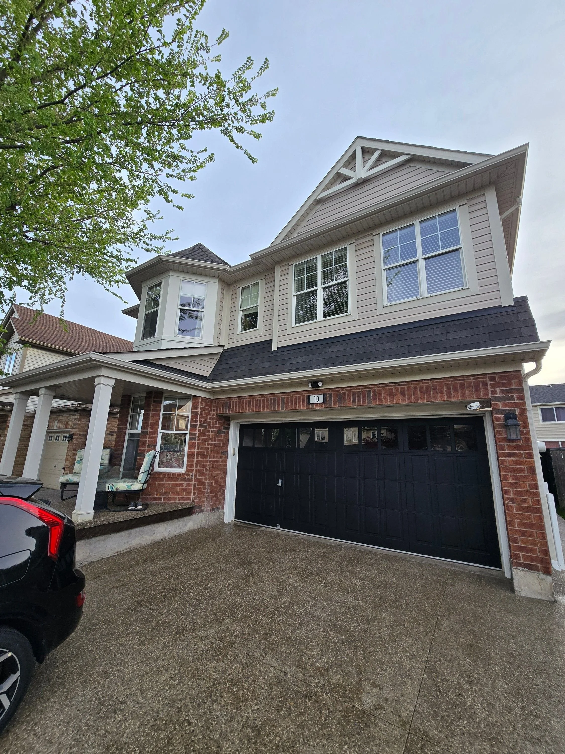 A two-story house with beige siding, black garage door, and brick accents. A black car is parked in the driveway, and porch furniture is visible near the front door. The house has multiple windows and a small porch with white columns. A tree with gre