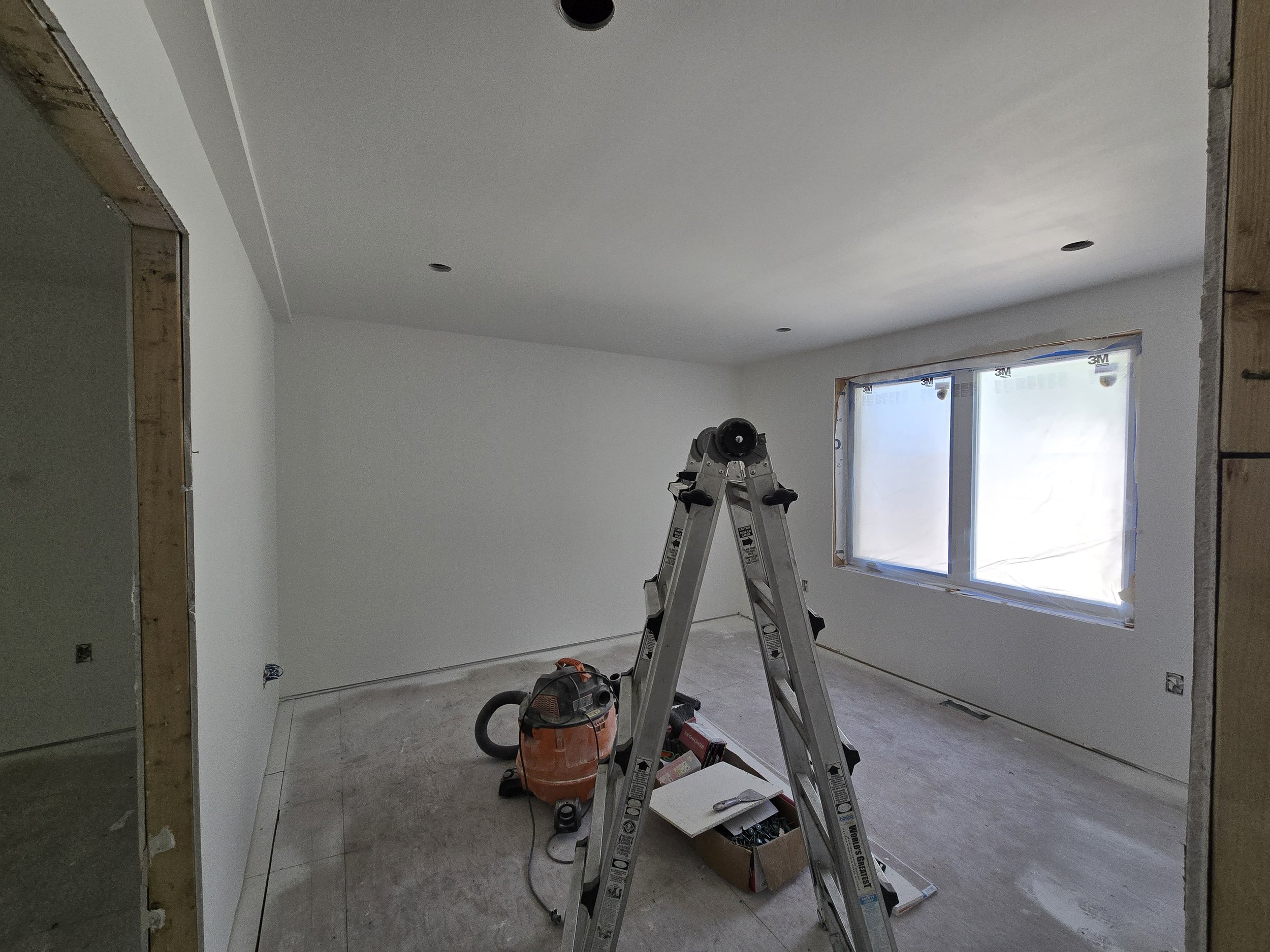A room under construction with a ladder, partially finished walls, a window with protective covering, and construction tools and materials on the floor.