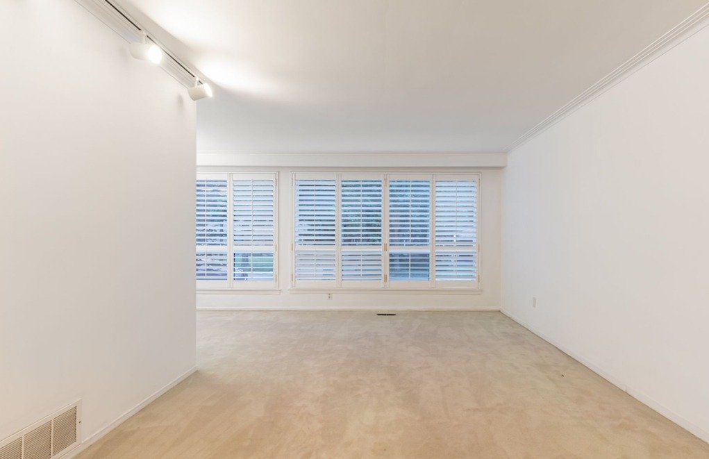 Empty room with white walls, light-colored carpet, and large windows with white blinds.