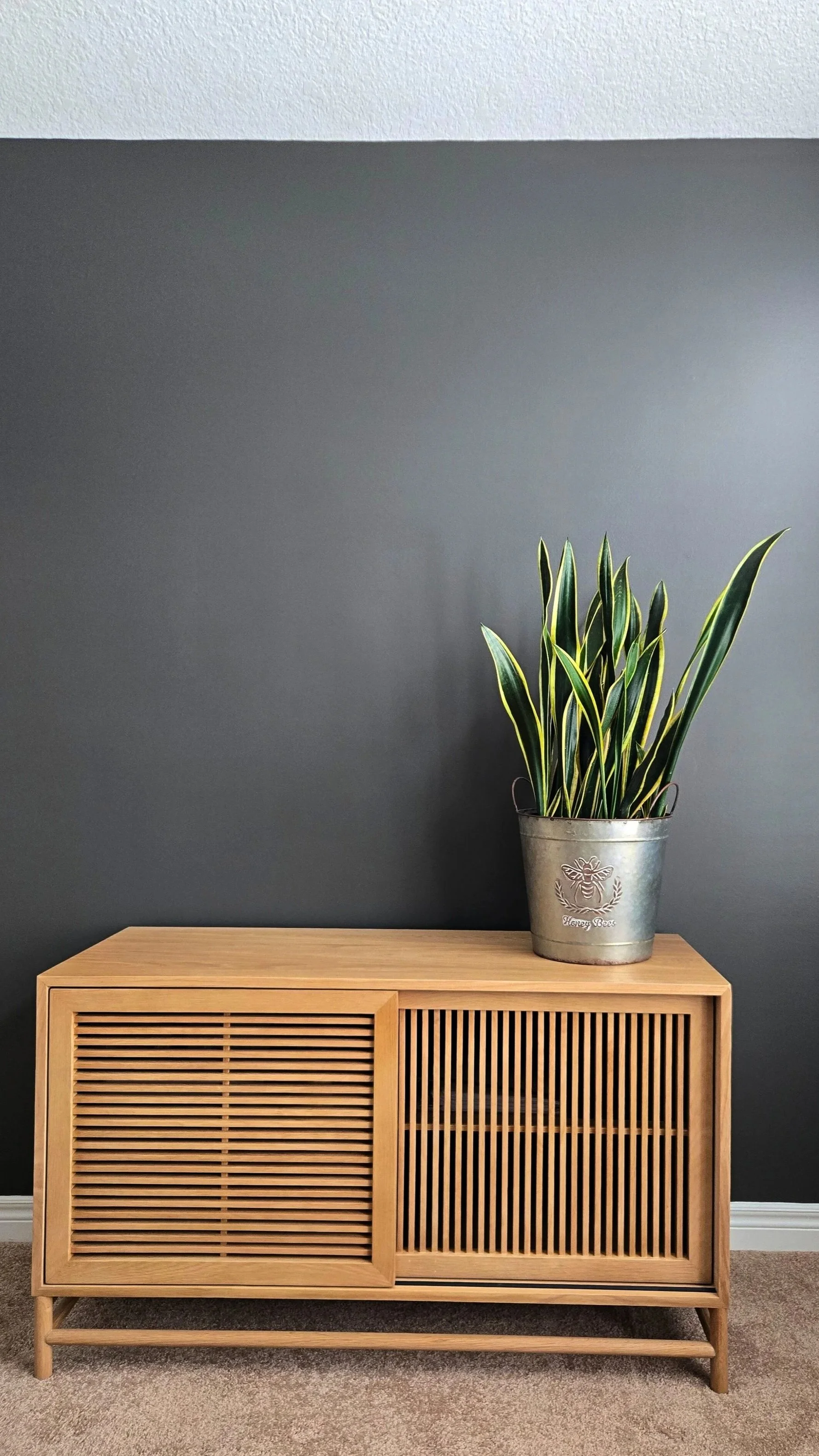 A wooden cabinet with slatted doors and a light wood finish, placed against a dark gray wall. On top of the cabinet is a metal pot with a decorative bee logo and the words 'Honey Bees' engraved on it, containing a green and yellow variegated plant.
