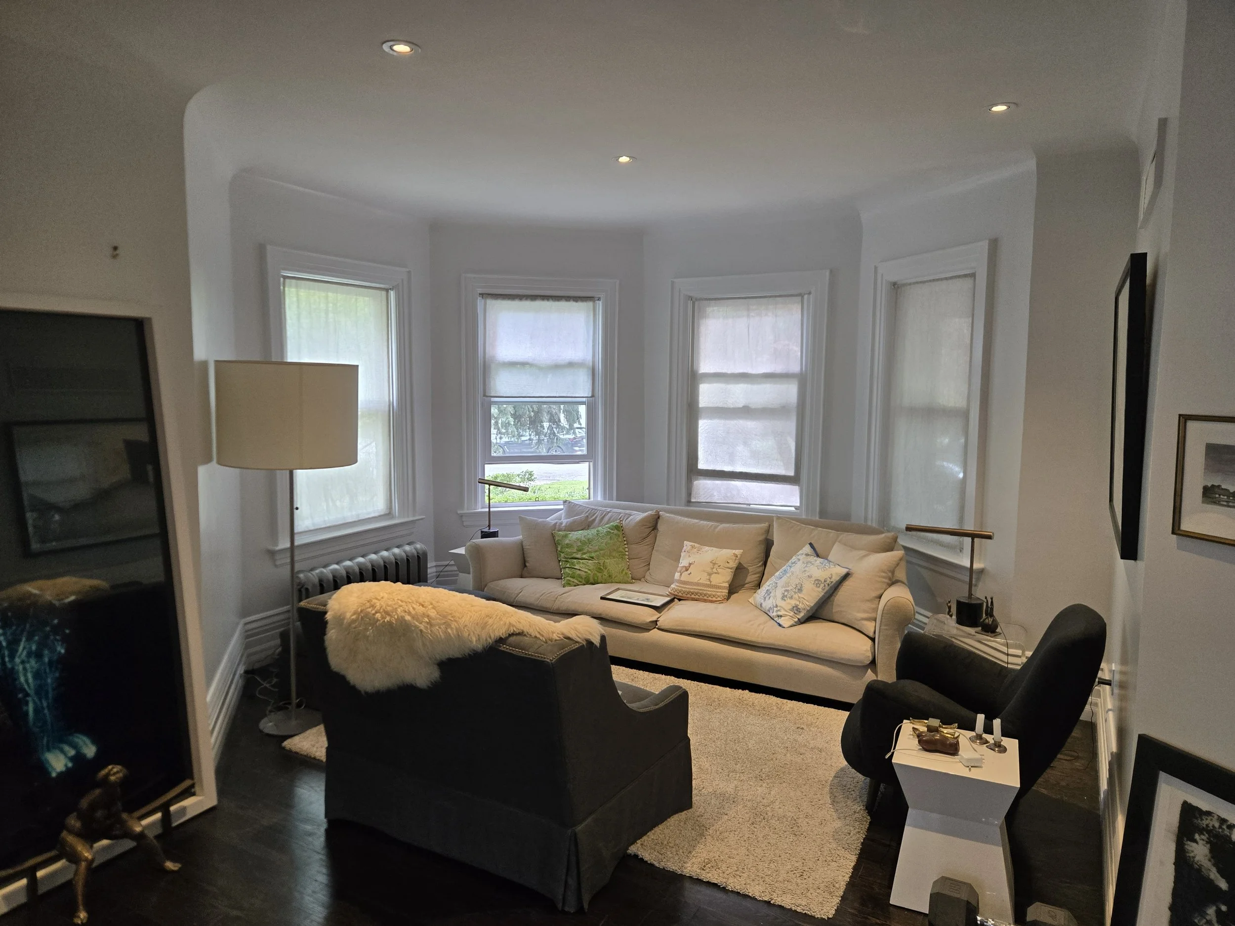 Living room with three large bay windows with white blinds, a beige couch with pillows, a black armchair with a furry blanket, a white side table with decor, and standing lamps, with a dark hardwood floor and a cream-colored area rug.