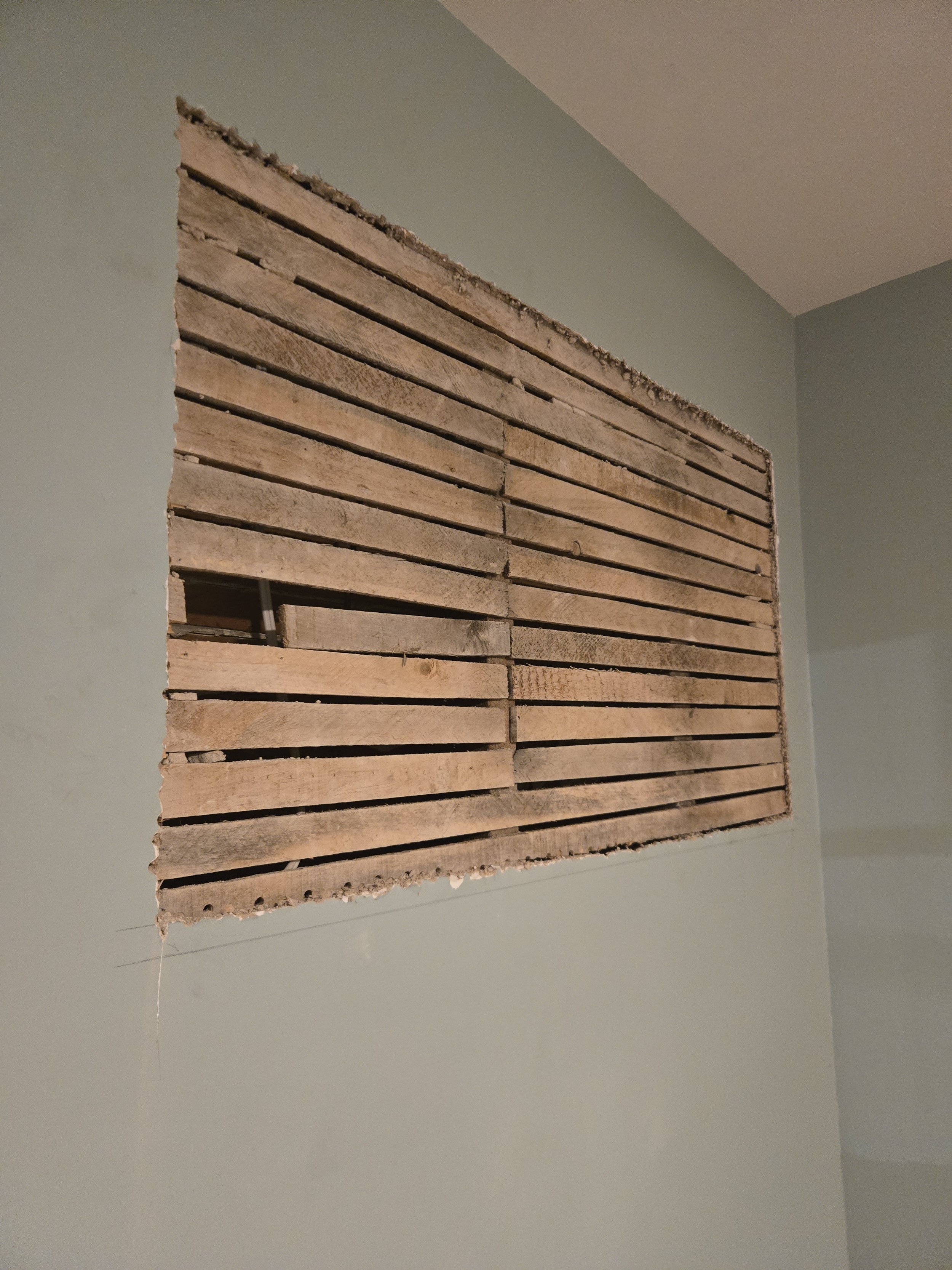A partially demolished wall with wooden slats and exposed drywall in a room with pale green walls.