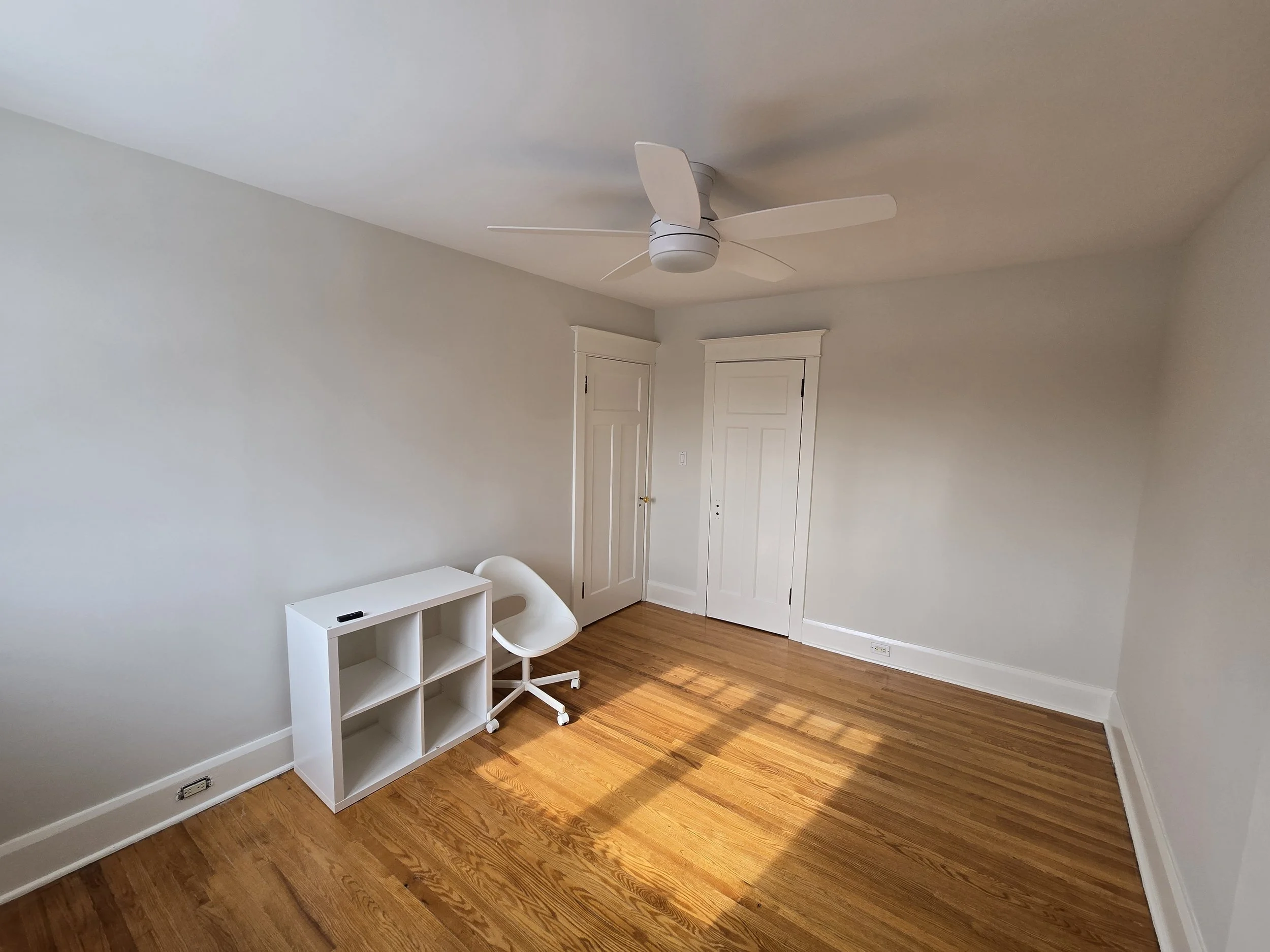A plain empty room with light-colored walls, hardwood floor, two white closed doors, a white desk, a white swivel chair, a ceiling fan with four blades, and an electrical outlet near the baseboard.
