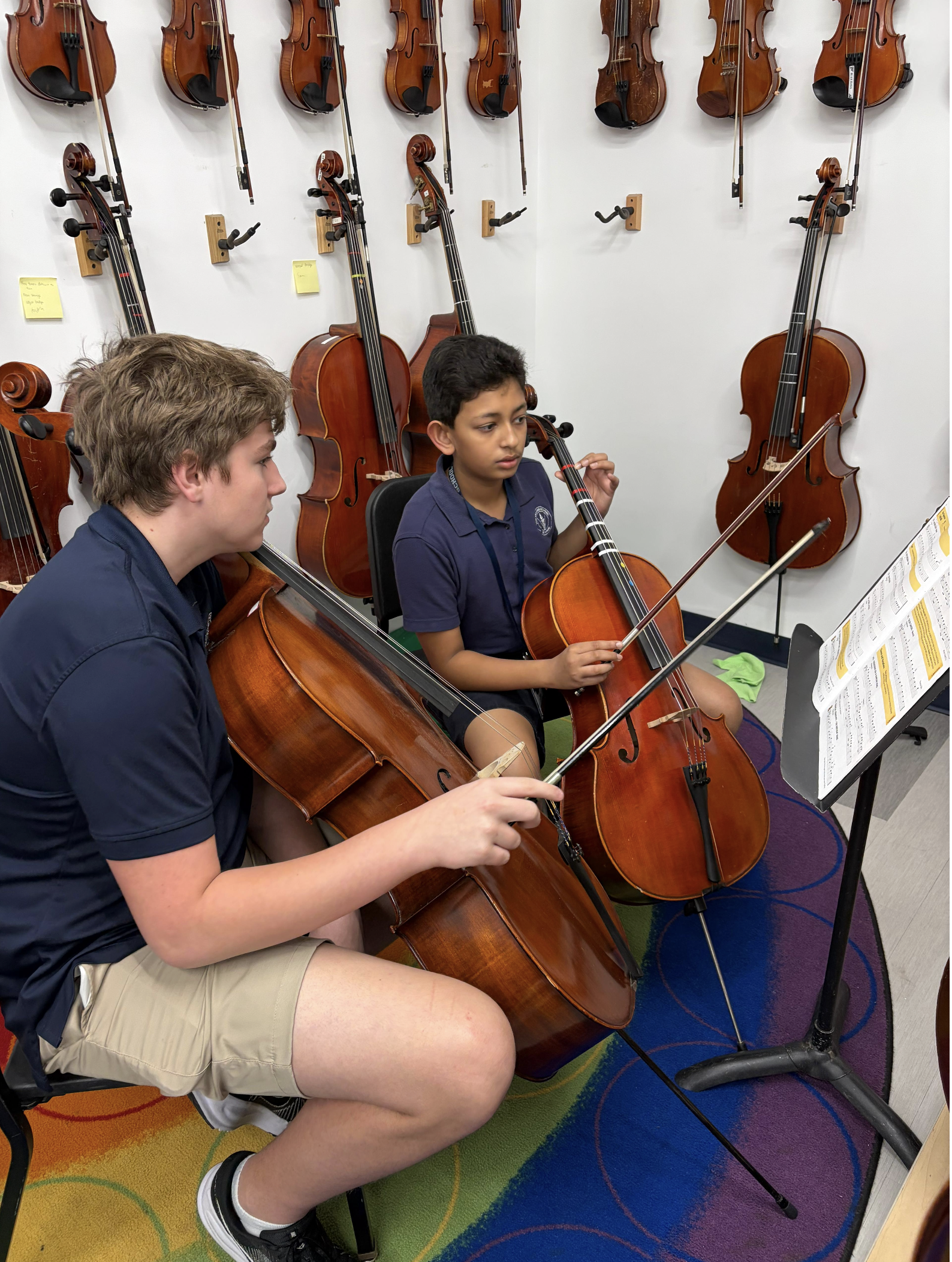 Two boys are sitting and playing cellos in a music classroom. The room has wall-mounted violins and violas. The boy in the foreground is wearing a navy shirt and beige shorts, while the boy behind him is wearing a blue polo shirt. Sheet music is on a stand in front of them.