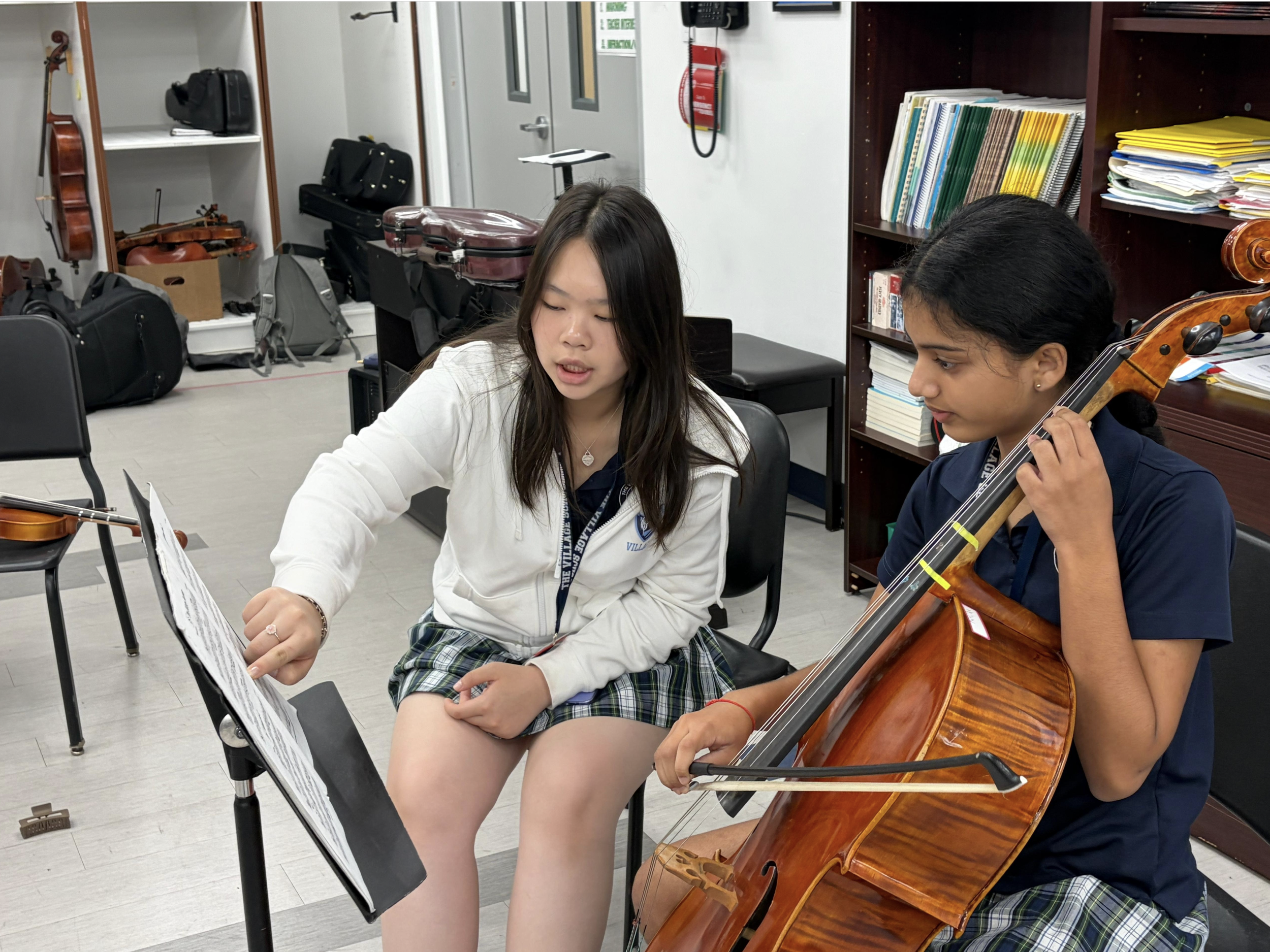 Two young women sitting side by side, one playing the cello and the other looking at sheet music in a classroom or music room setting.