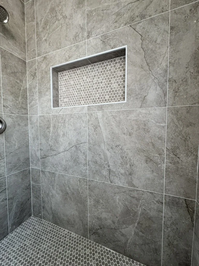 A tiled shower with grey marble tiles, a built-in niche with small hexagon tiles, and a drain in the corner.